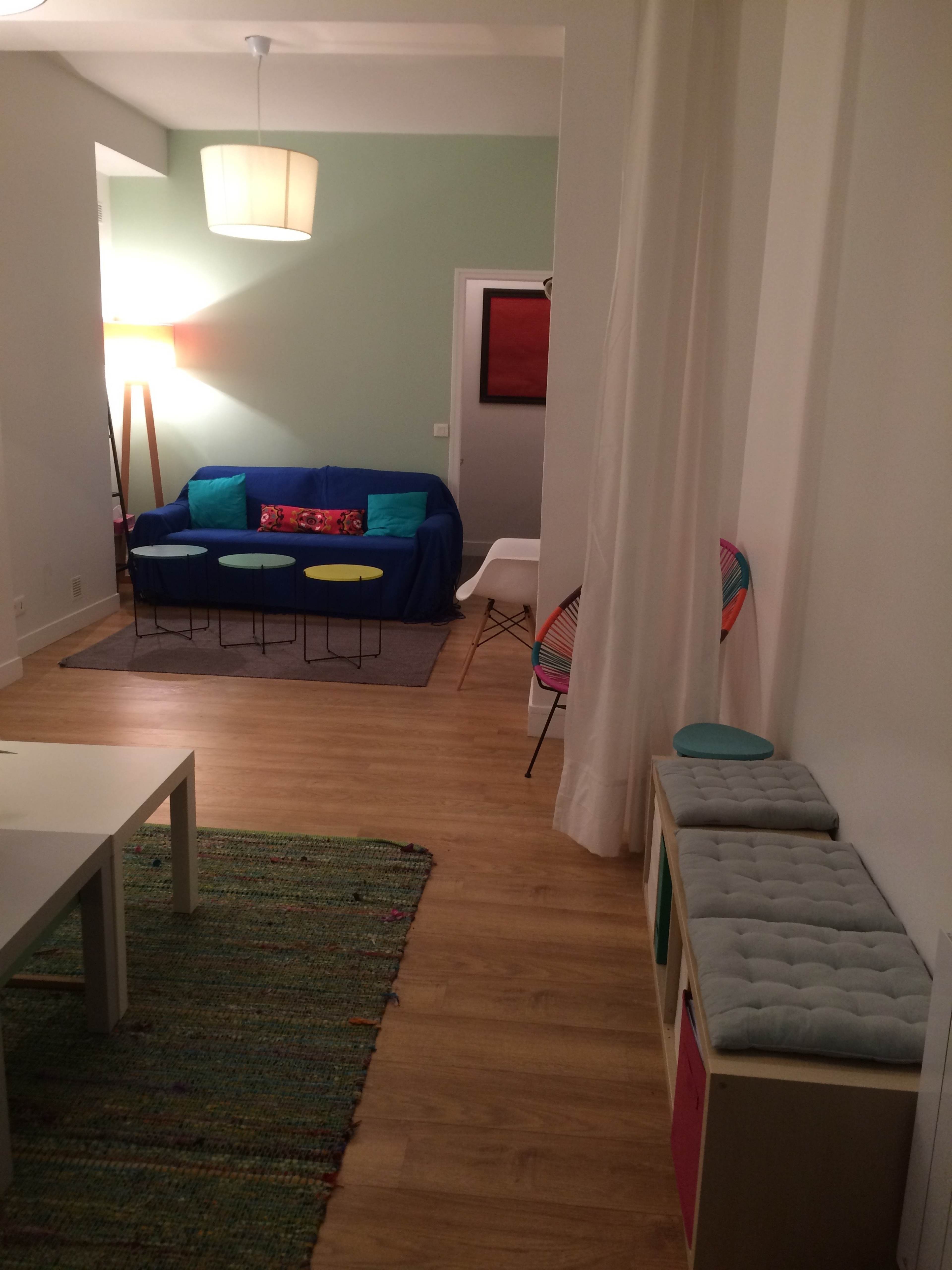 The image shows a living space with a blue sofa, a small coffee table, colorful stools, and a carpet, leading to an entryway with decor and furniture.