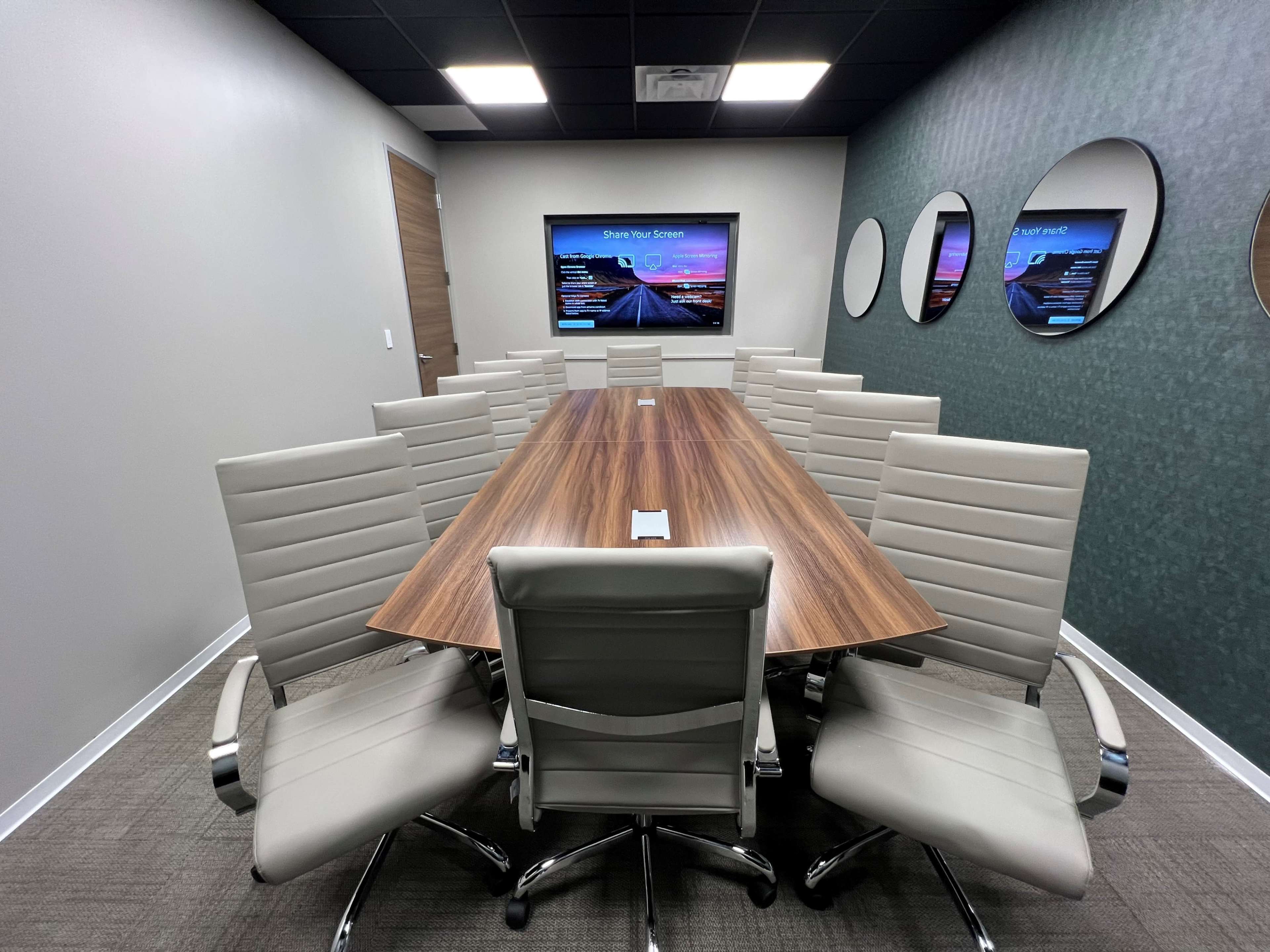 The image shows a modern conference room with a long wooden table, surrounded by twelve light-colored chairs, and two screens displaying information on the walls.