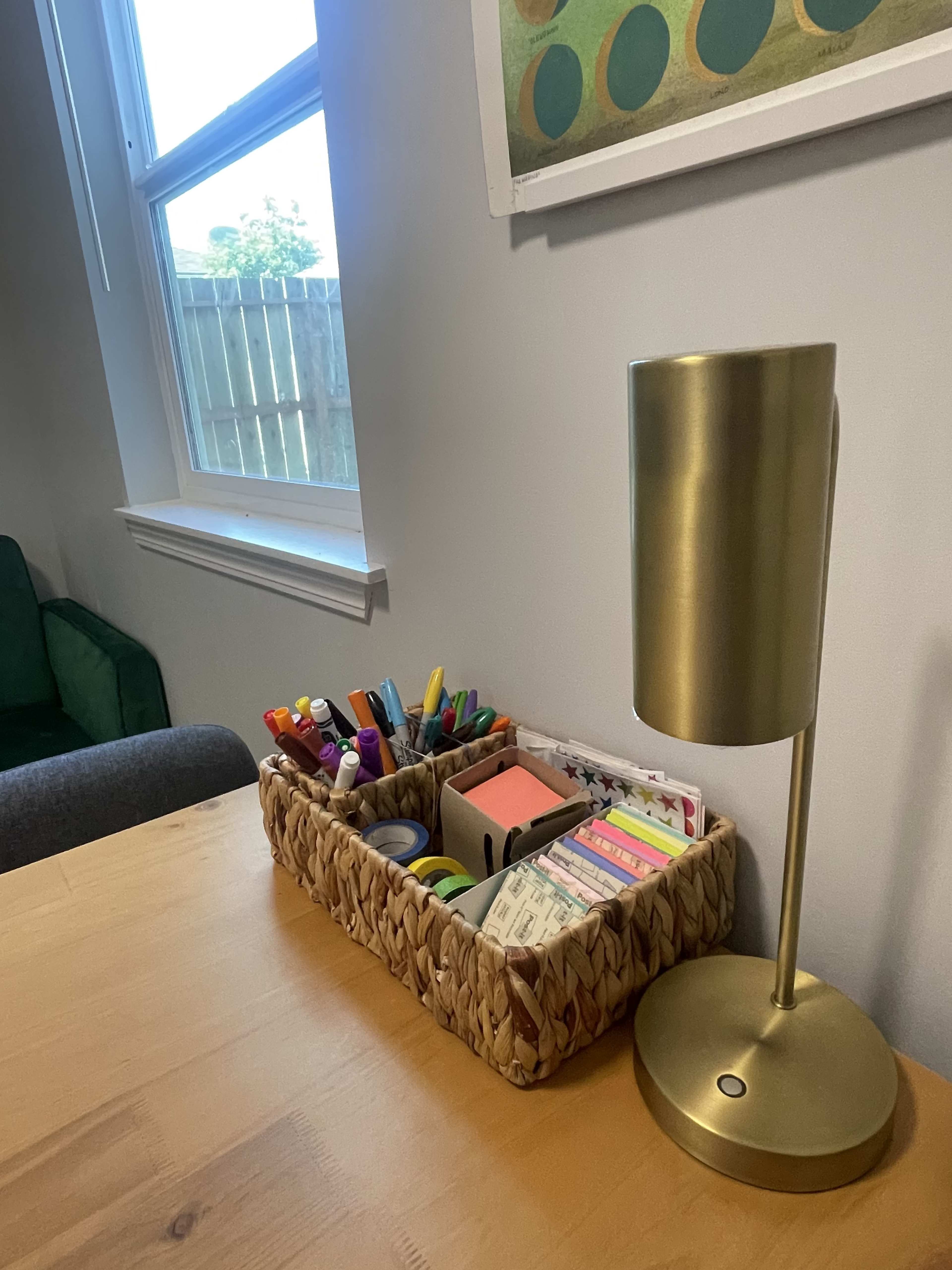 A wicker basket filled with various stationery items sits on a wooden table next to a gold lamp in a room with a window.