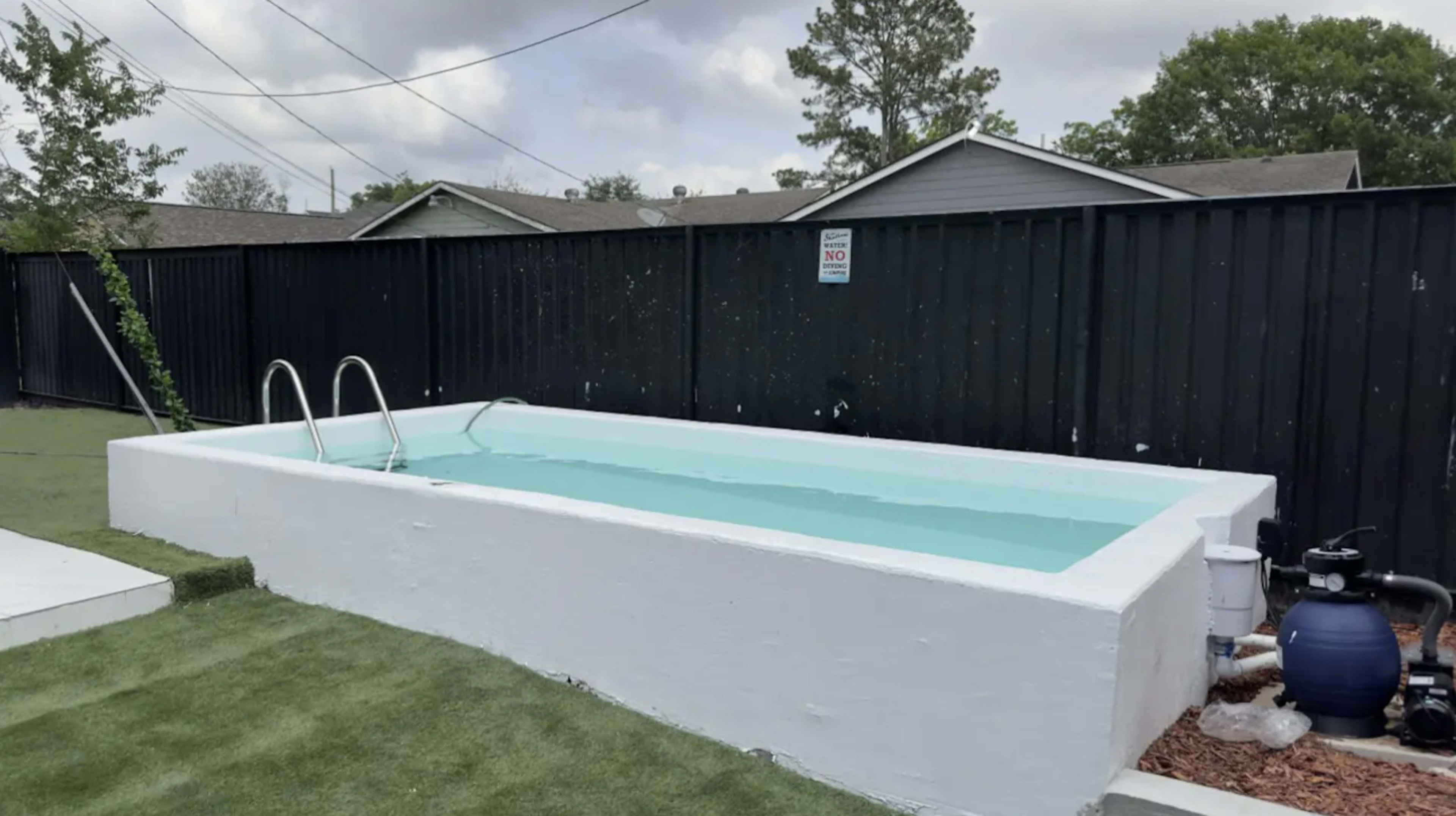 A rectangular above-ground swimming pool with clear water surrounded by artificial grass and a black fence in the background.