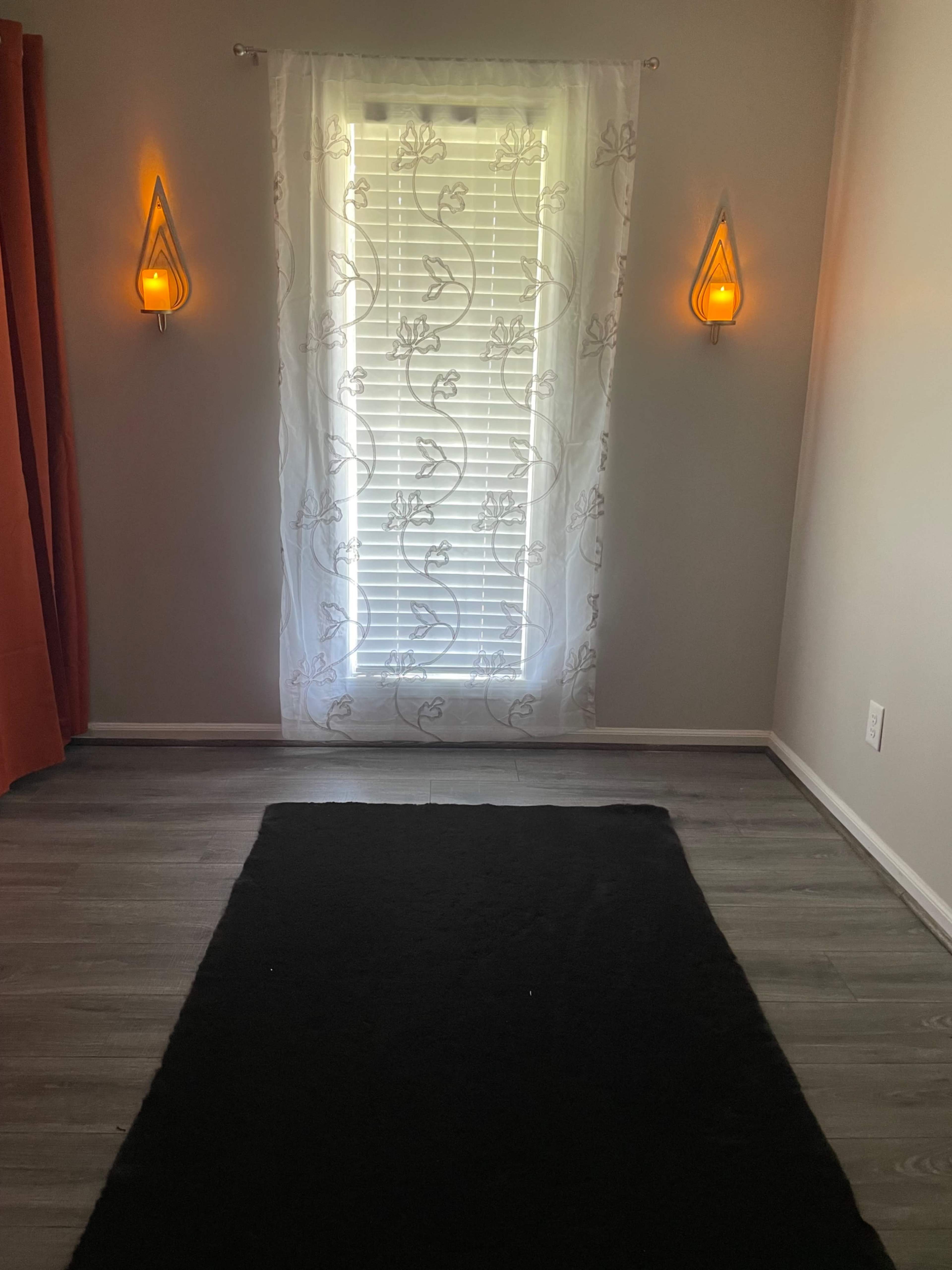 The image shows a room with a window covered by a sheer curtain, flanked by two wall sconces, and features a dark area rug on a gray floor.