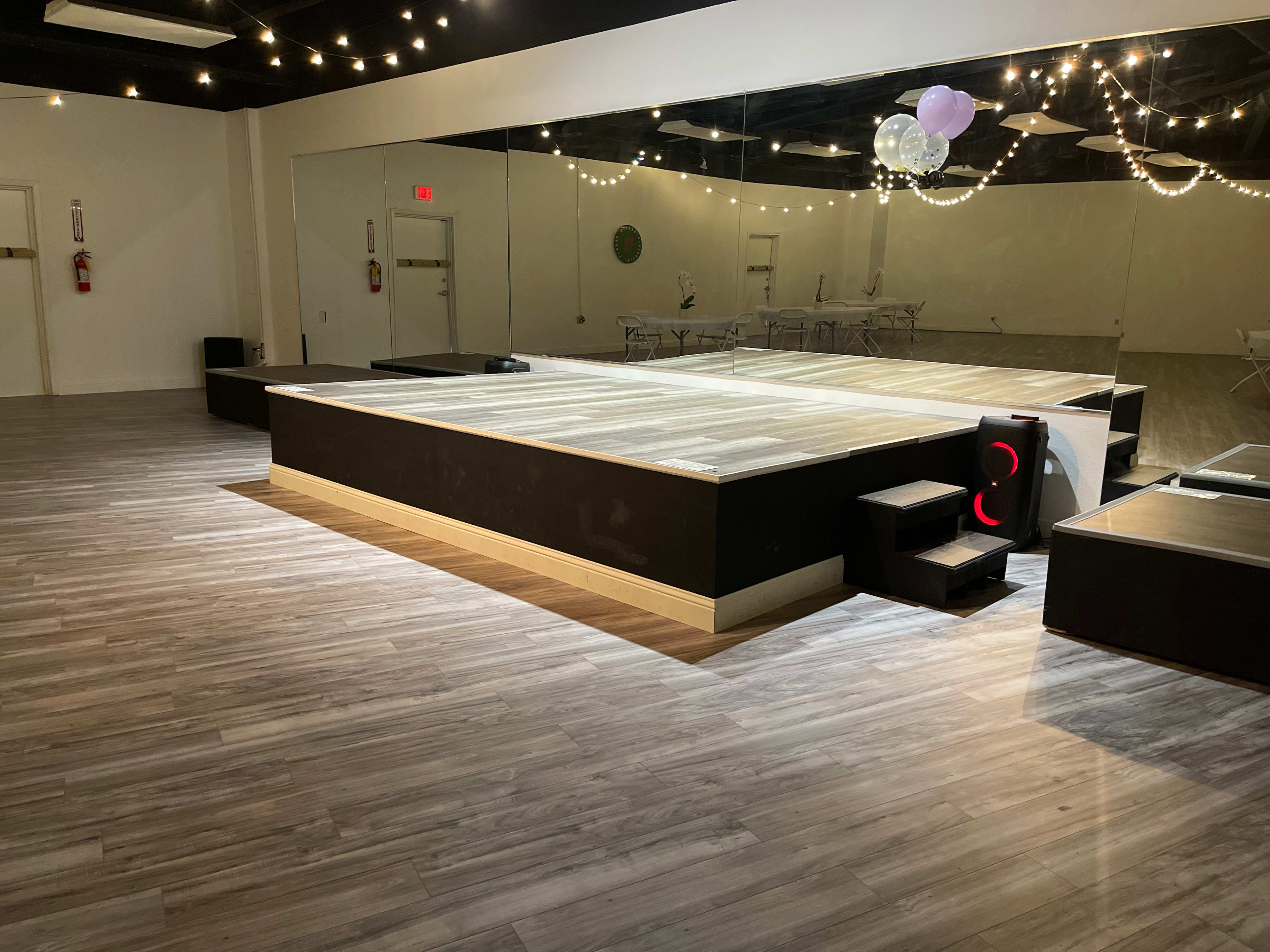 A large, elevated wooden platform surrounded by smooth, light-colored flooring in a spacious, well-lit room decorated with string lights.