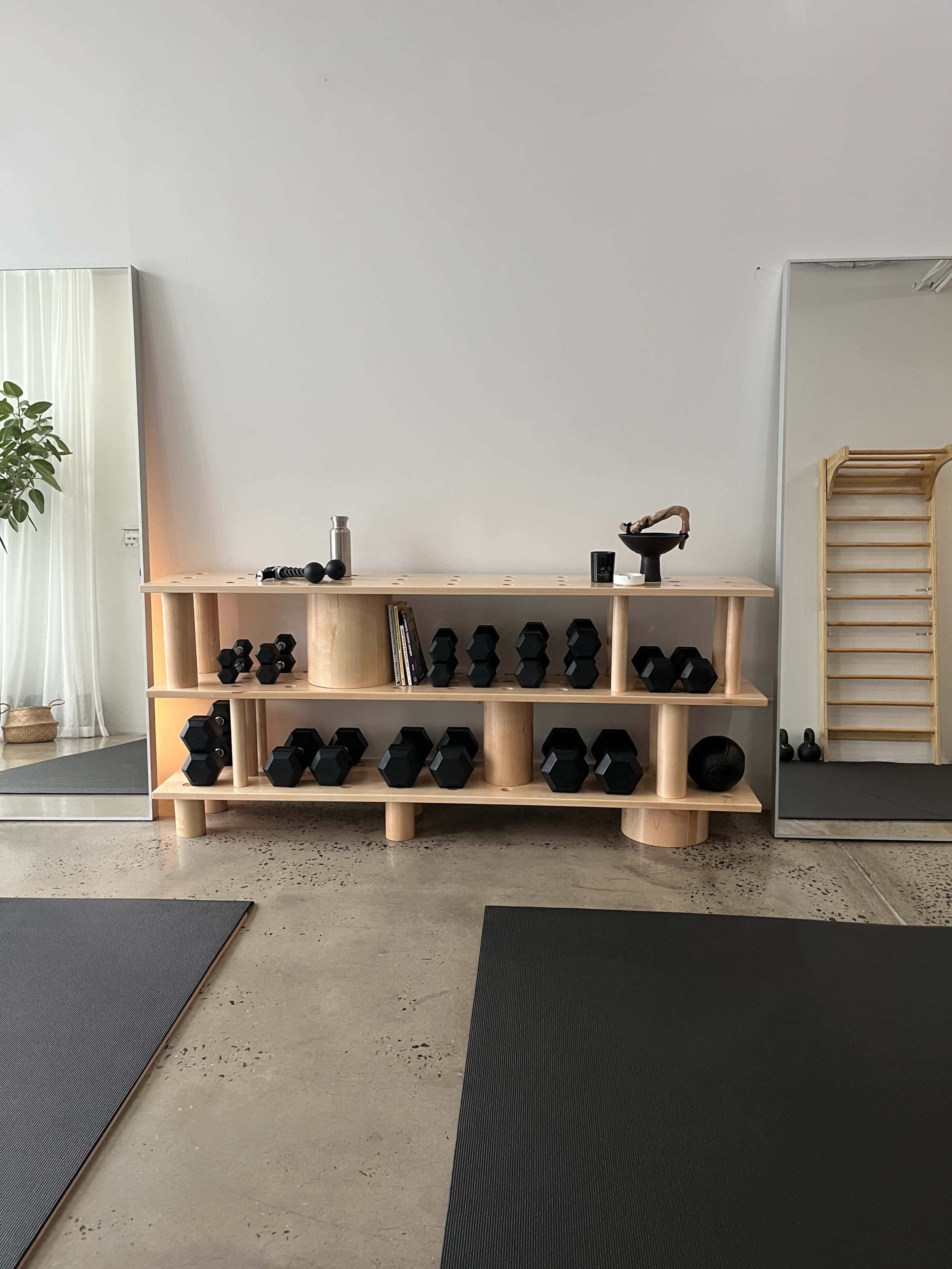 The image shows a minimalist fitness studio with a wooden shelf displaying various black dumbbells and a large mirror reflecting the space.