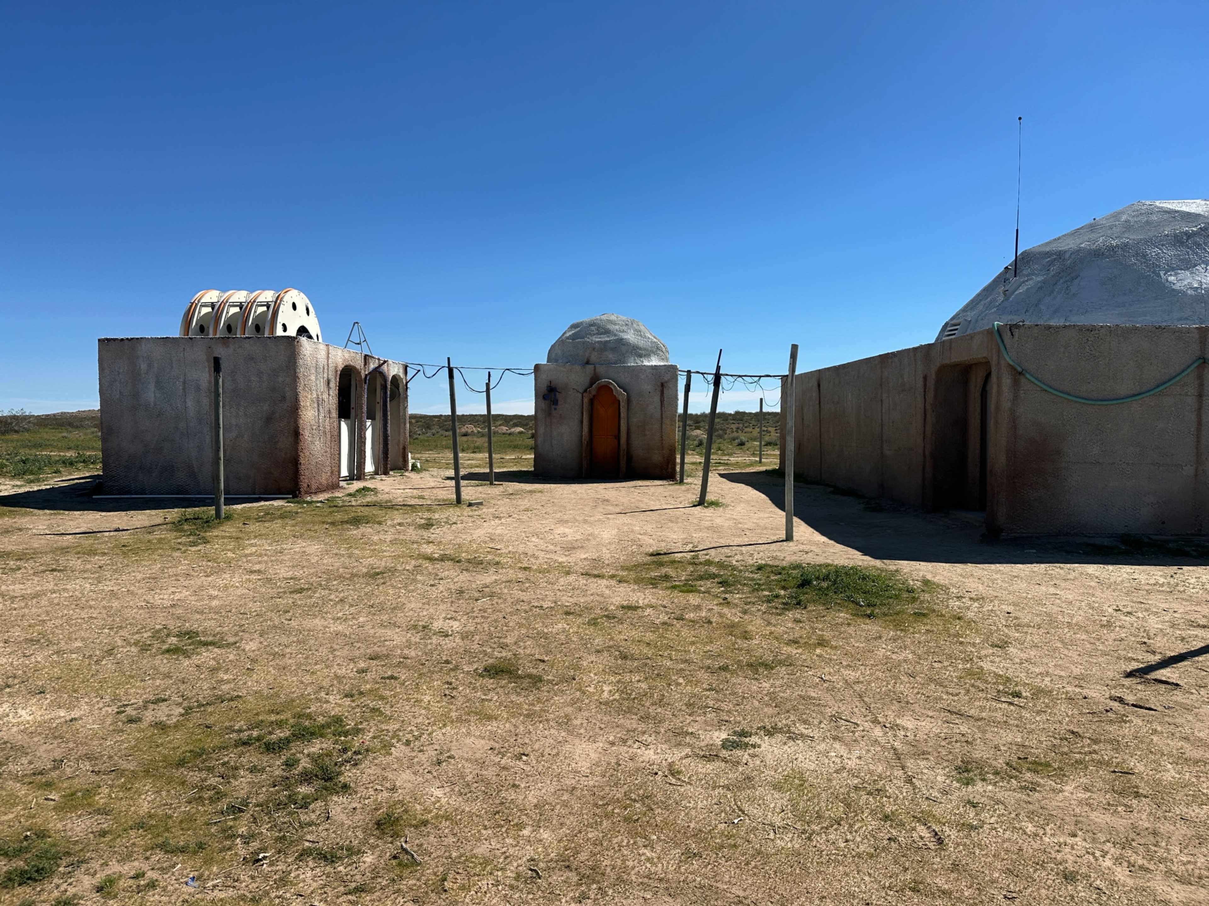 The image shows a barren landscape with three unique structures, two of which have domed roofs, and a series of clotheslines in between.