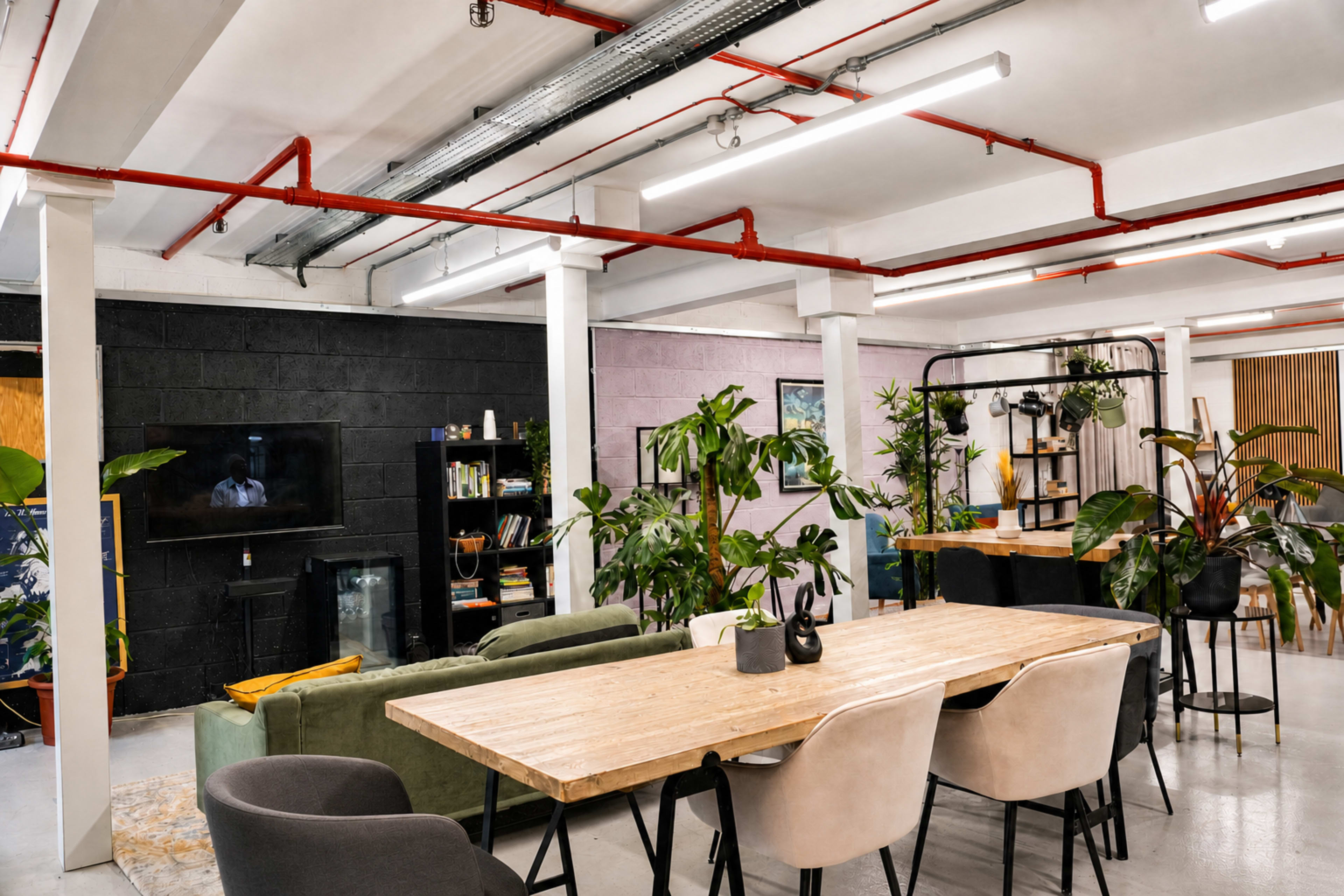 A modern workspace features a long wooden table surrounded by chairs, plants, and distinct areas for work and relaxation.