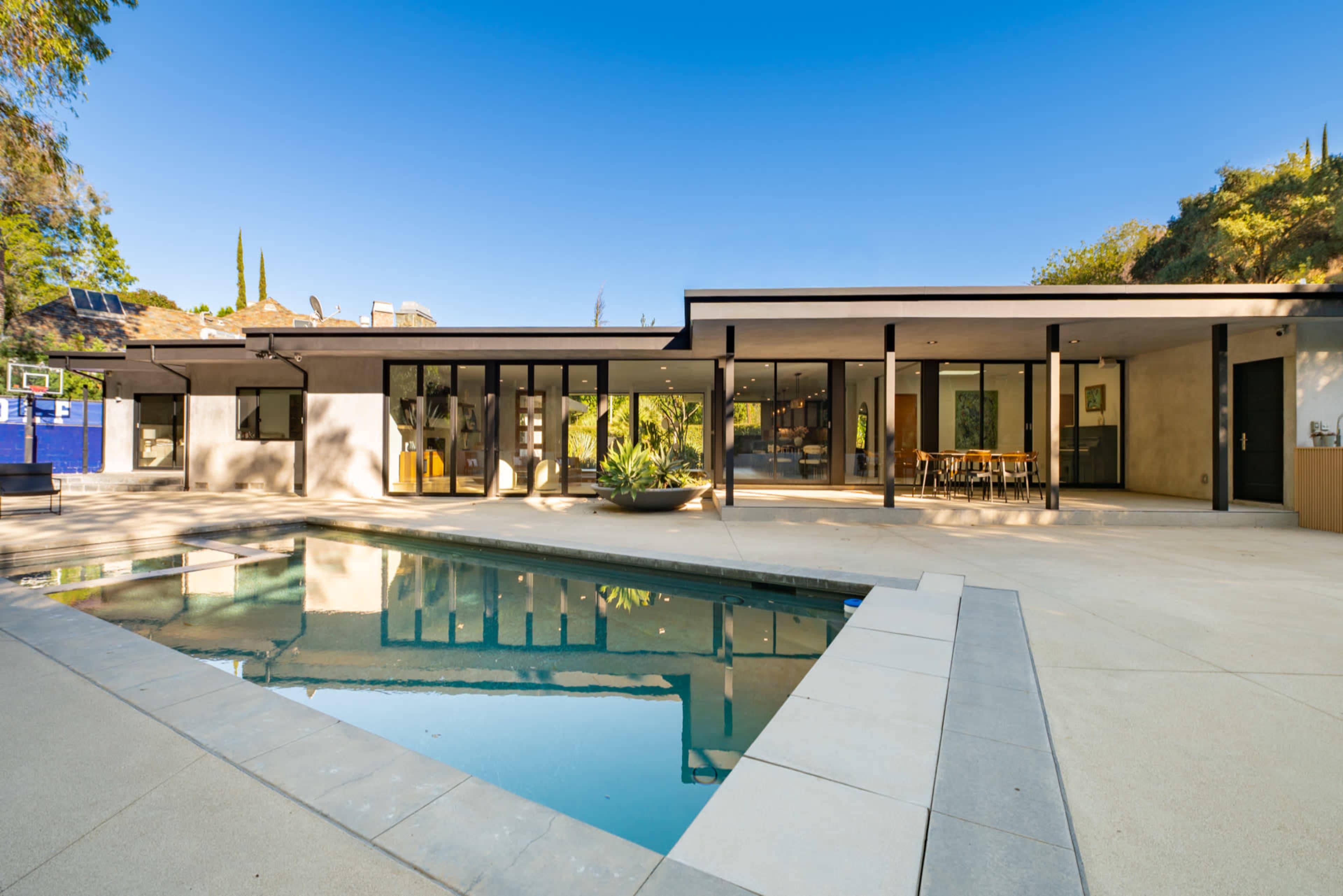 The image shows a modern one-story house with large glass doors opening into a backyard featuring a swimming pool surrounded by concrete and greenery.