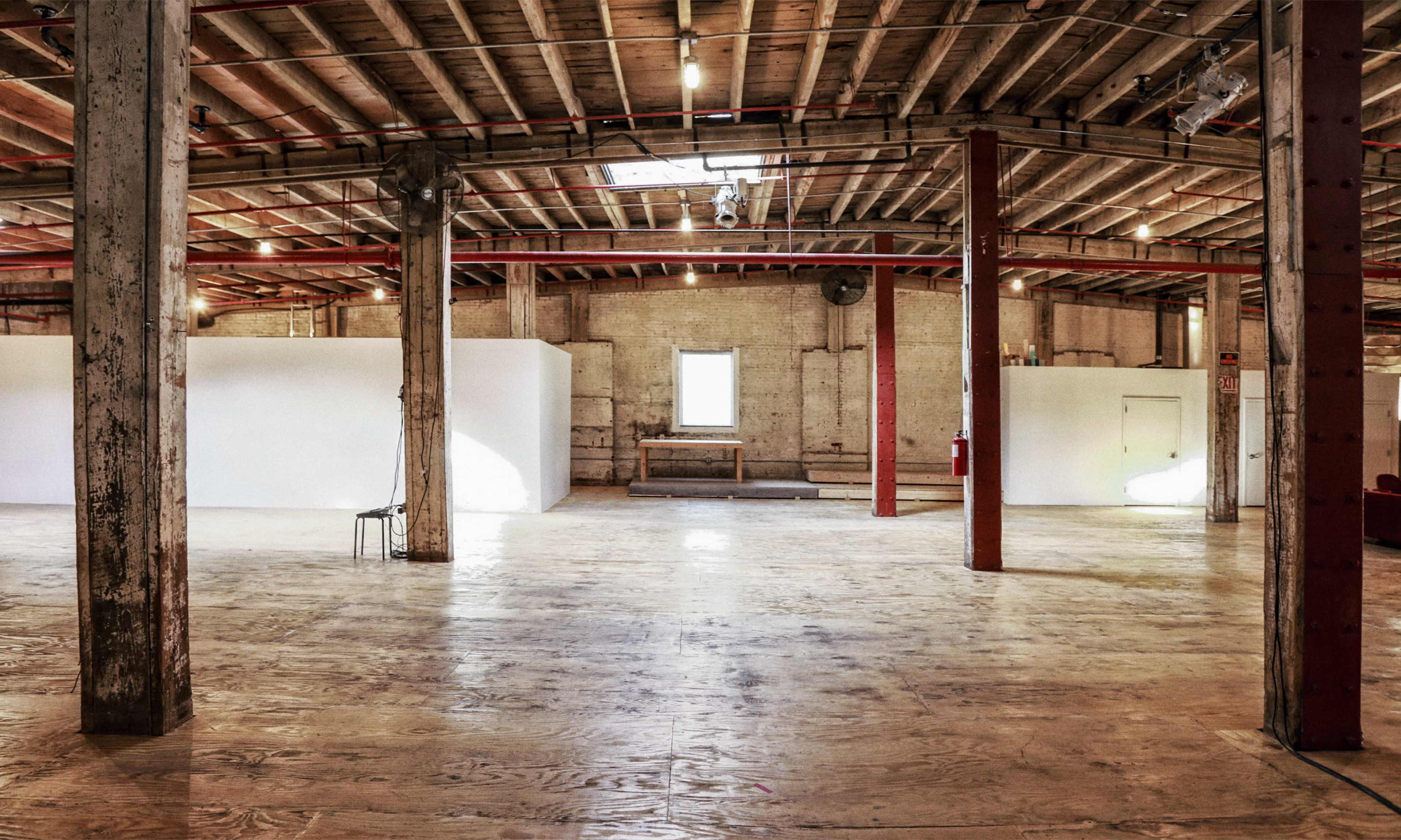 The image shows a spacious, empty industrial interior with wooden flooring and exposed beams, featuring white walls and a single window in the background.