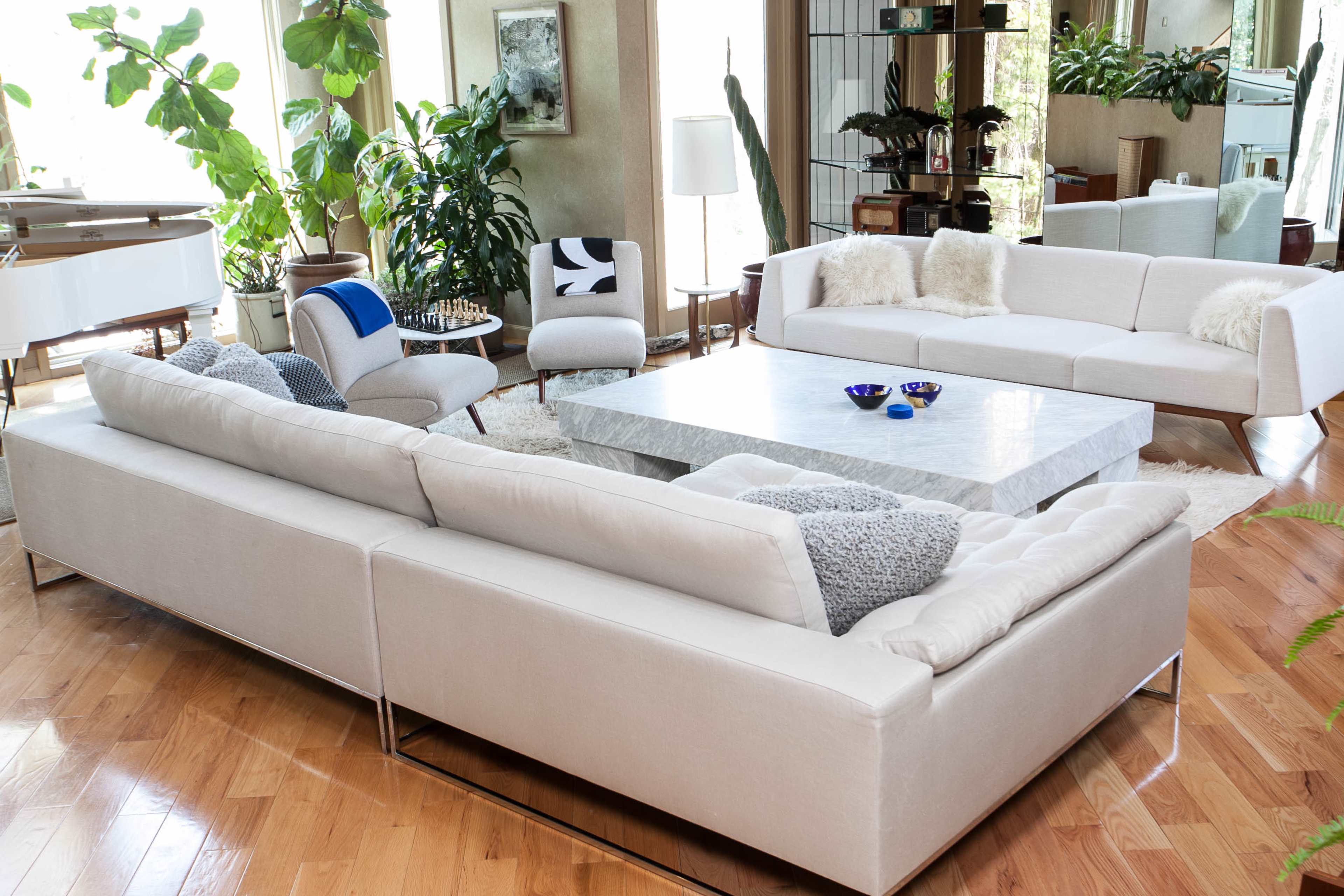 The living room features a spacious layout with a large stone coffee table surrounded by a light-colored sectional sofa and contemporary chairs, all set among various indoor plants.