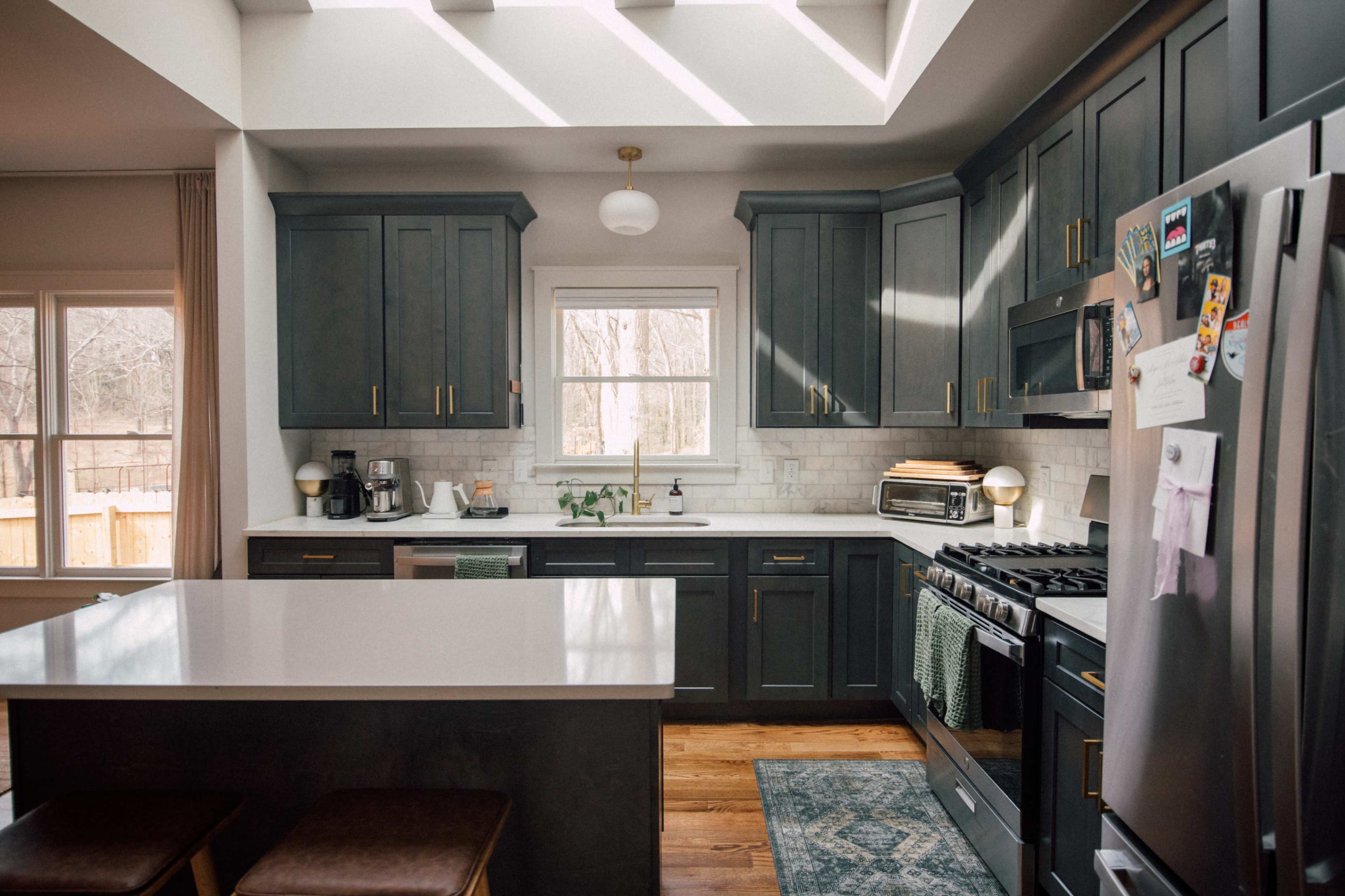 The kitchen features dark cabinetry, a central island with seating, and stainless steel appliances, illuminated by natural light from skylights.