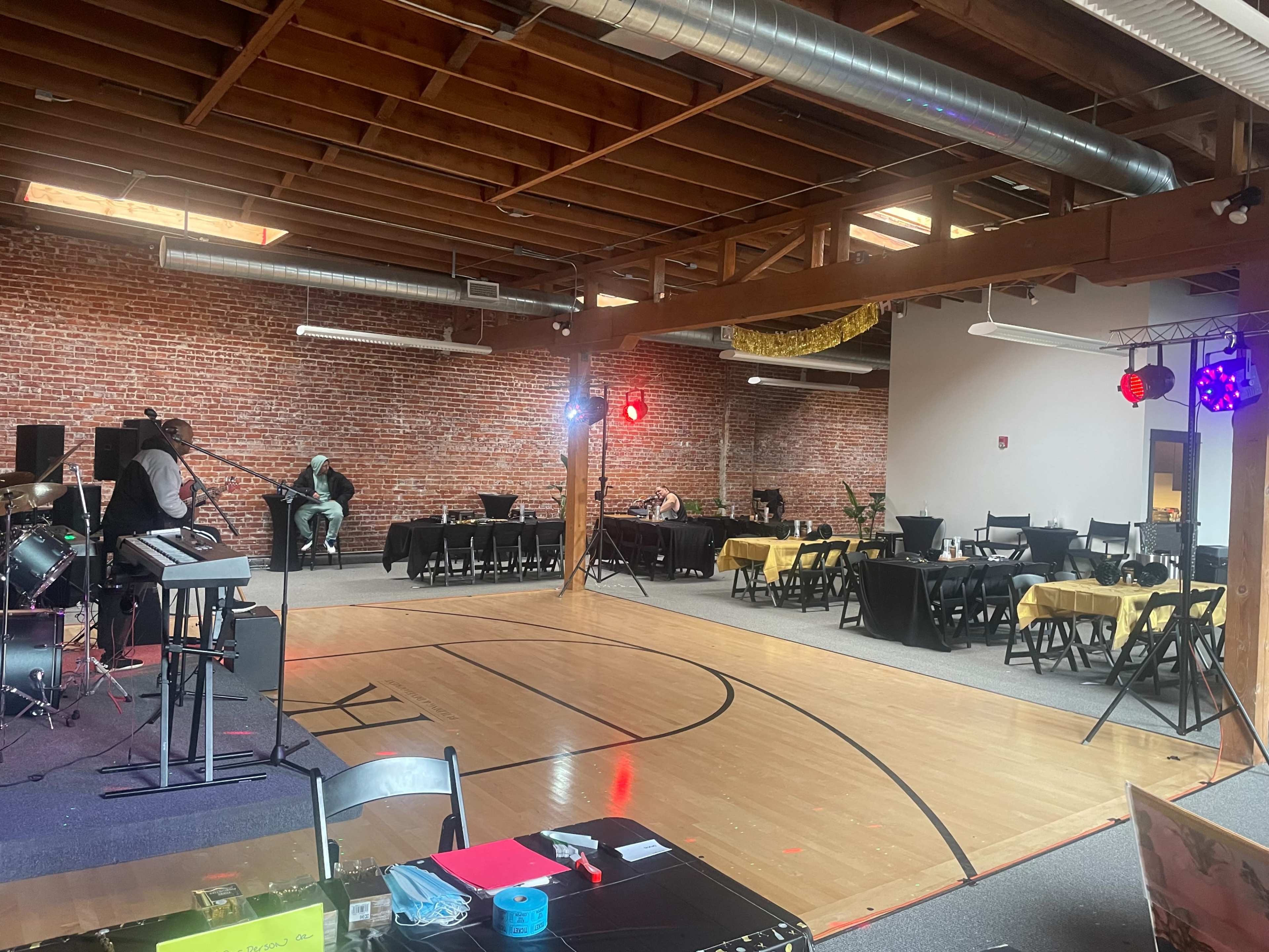 The image shows an empty event space with hardwood flooring, black tables set for dining, and a small stage area with musical equipment.