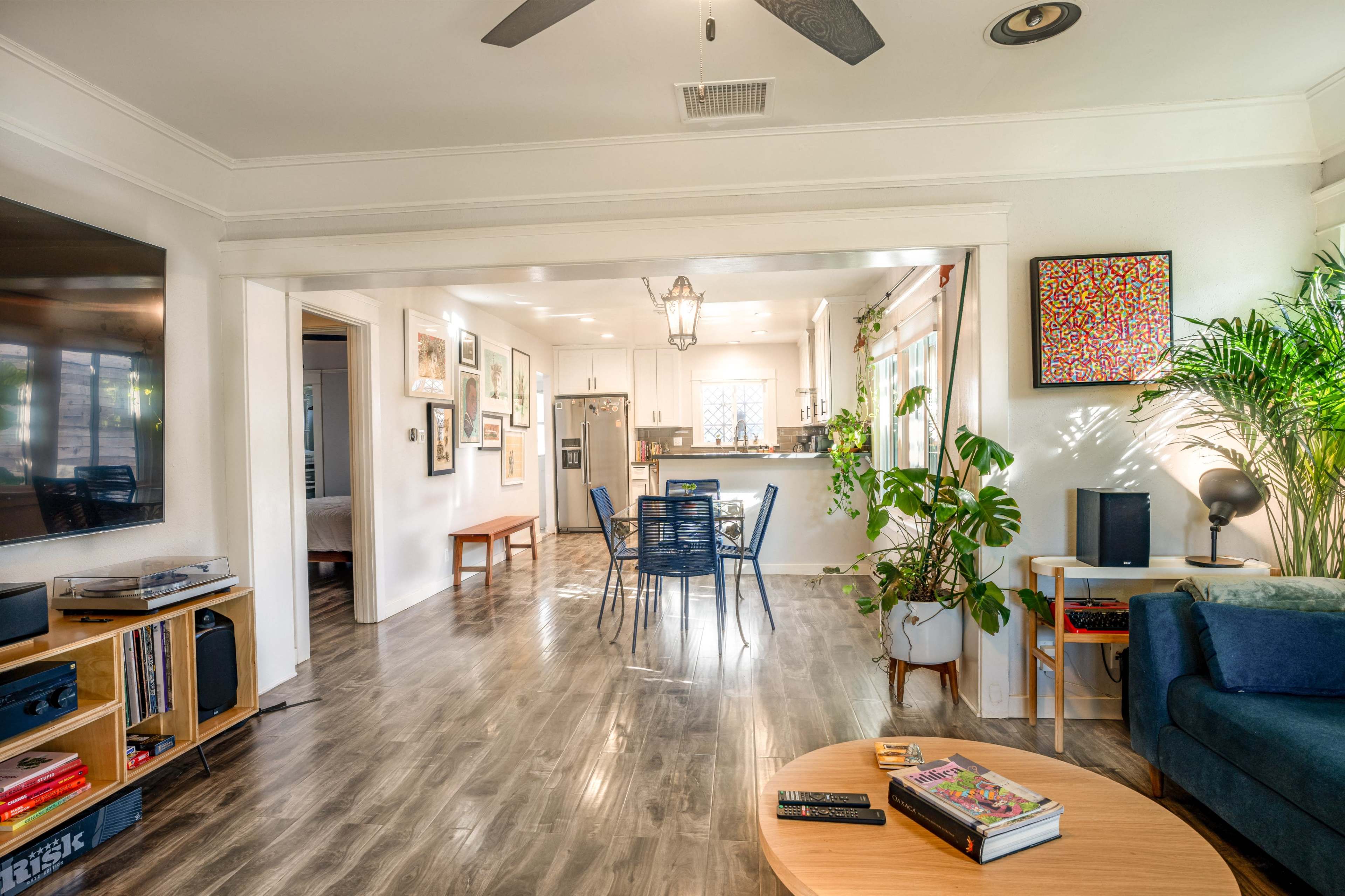 The image shows a modern, open-concept living space featuring a dining area with a blue sofa, a wooden coffee table, and various decorative plants.