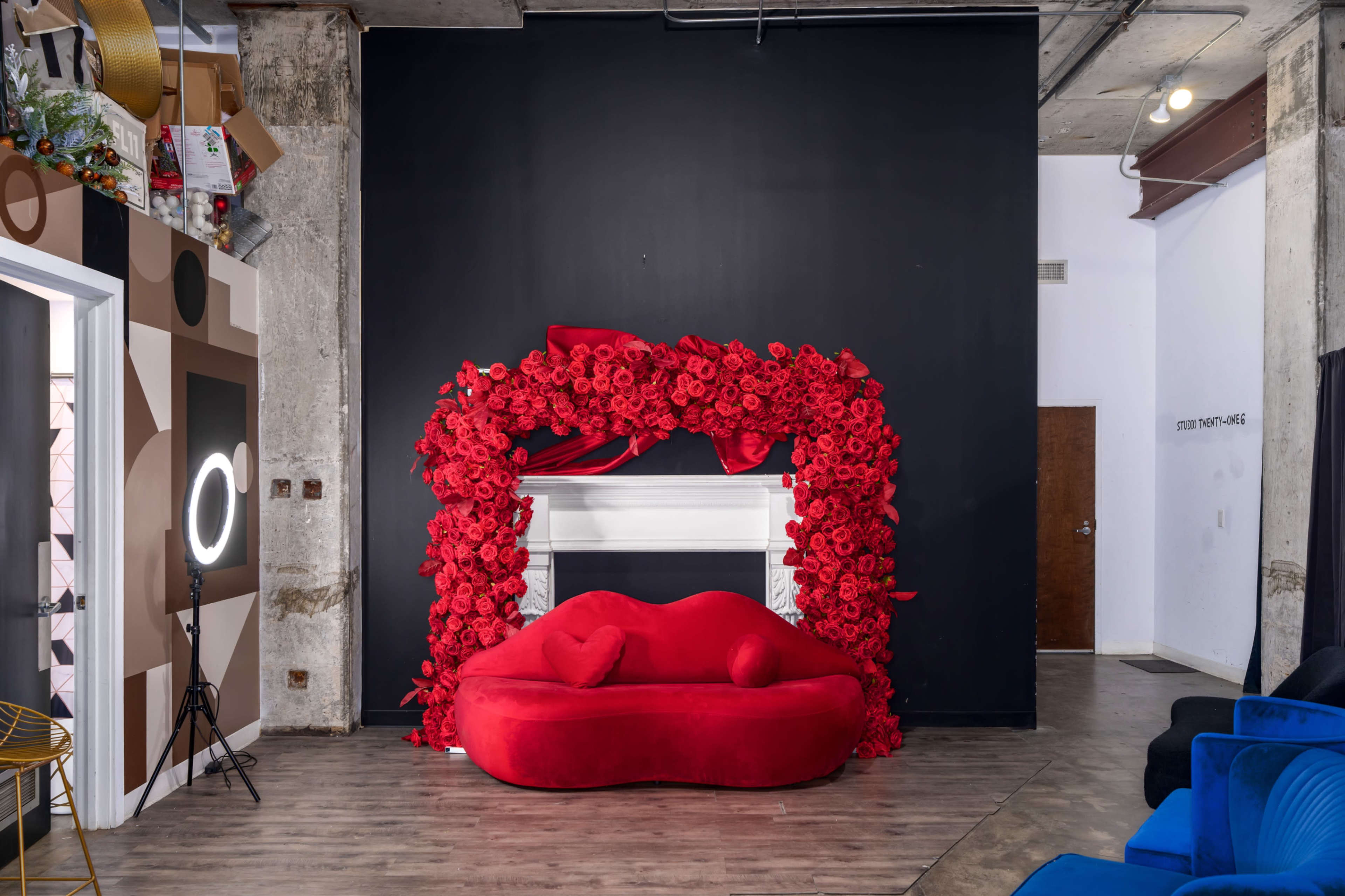 A red sofa is placed in front of a black wall adorned with an arch of red flowers, framed by a white mantelpiece.