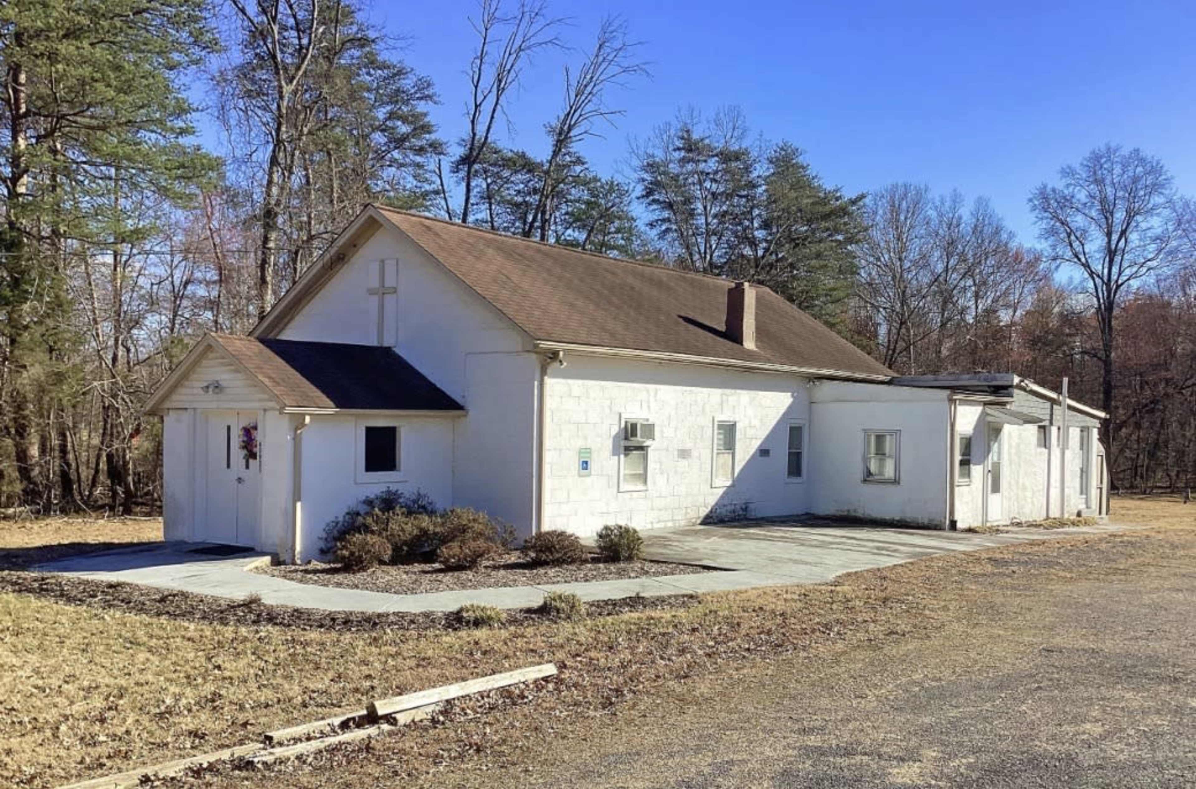 The image shows a simple white church building with a cross on its front, surrounded by a wooded area and a gravel pathway.