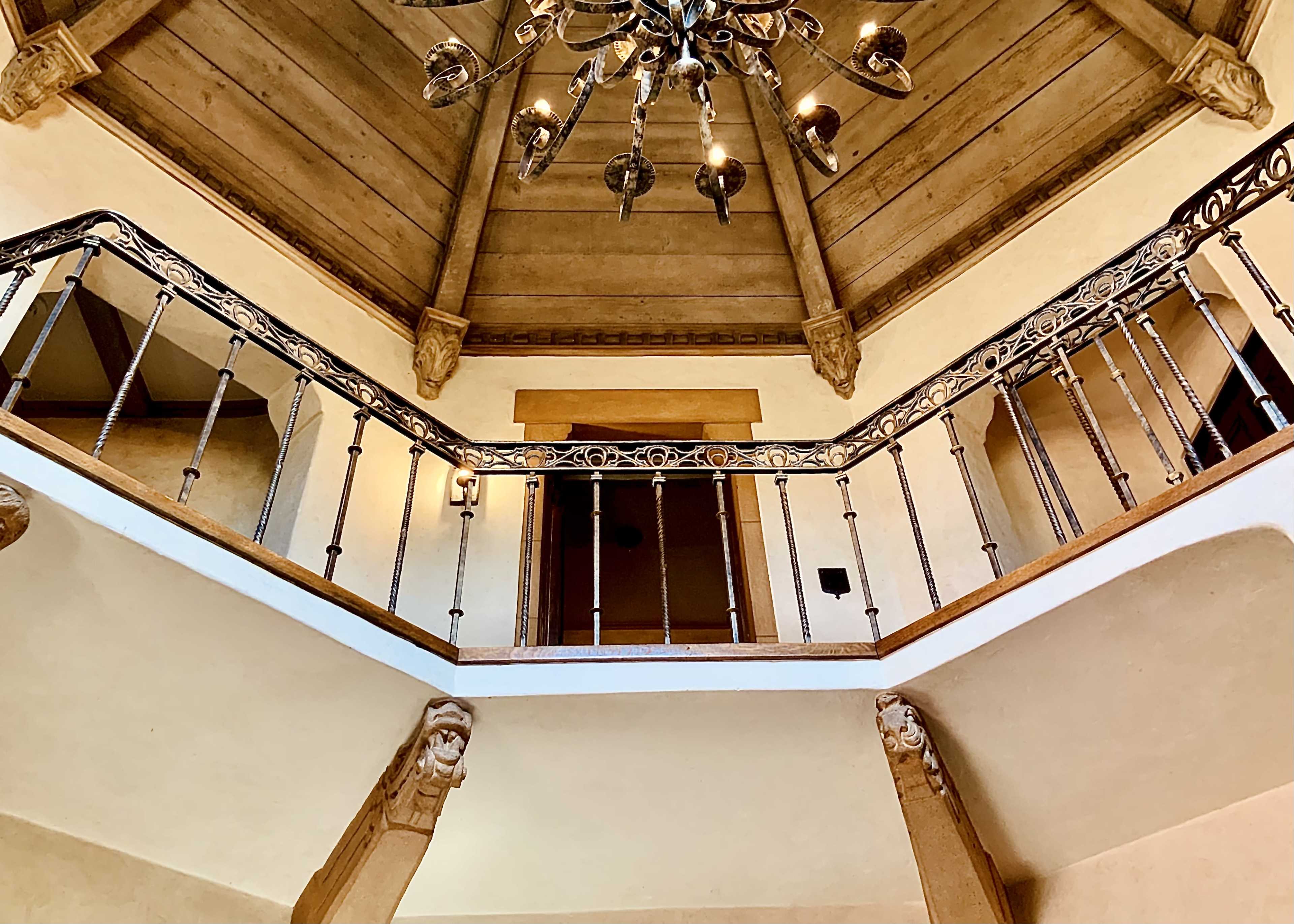 The image shows an ornate staircase with a wrought iron railing, leading up to a vaulted ceiling with a chandelier in a well-lit hallway.