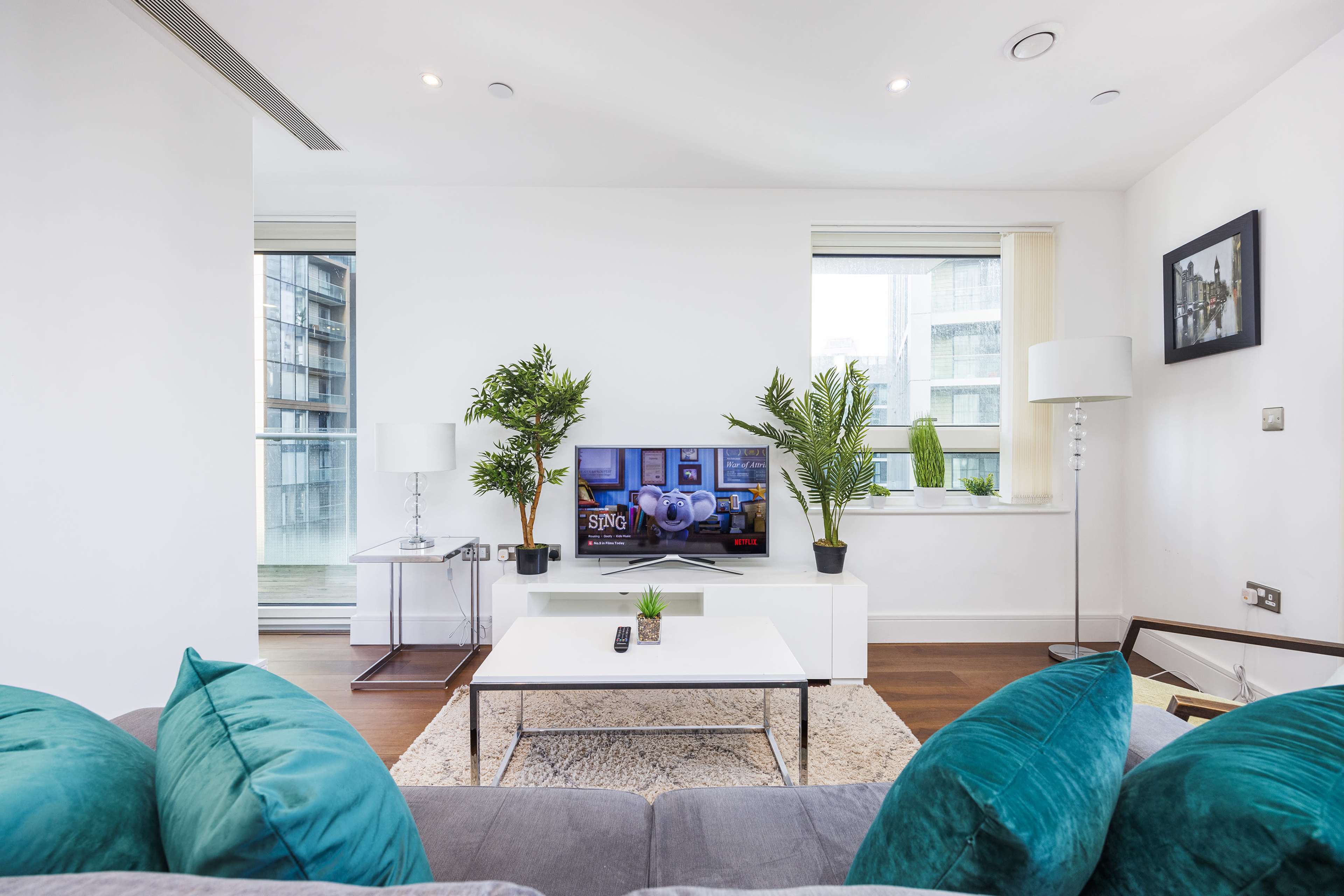 A modern living room features a gray sofa with teal cushions, a white coffee table, and a television on a stand, surrounded by potted plants.