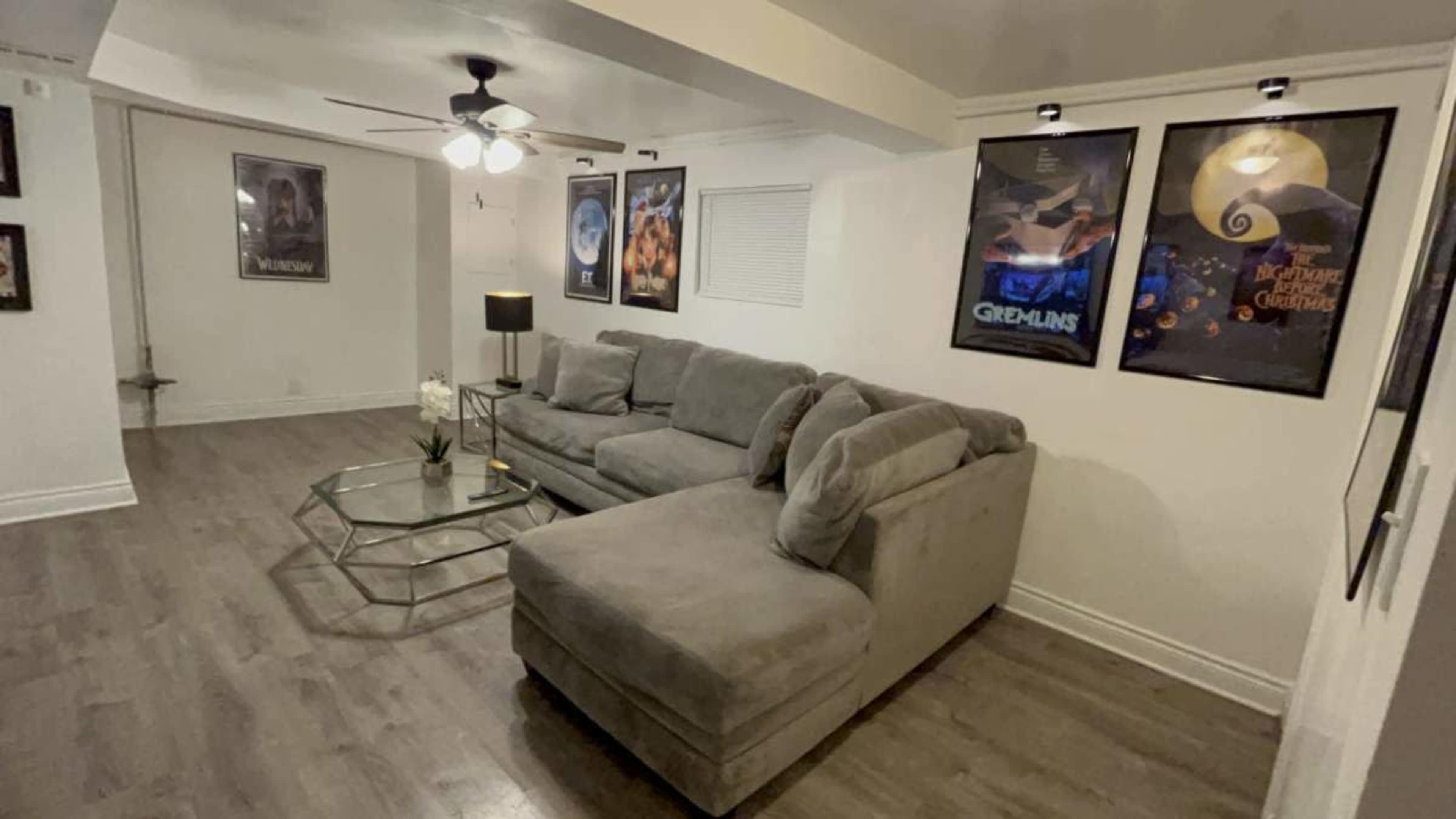 The image shows a modern living room featuring a gray sectional sofa, a glass coffee table, and several movie posters hanging on the walls.