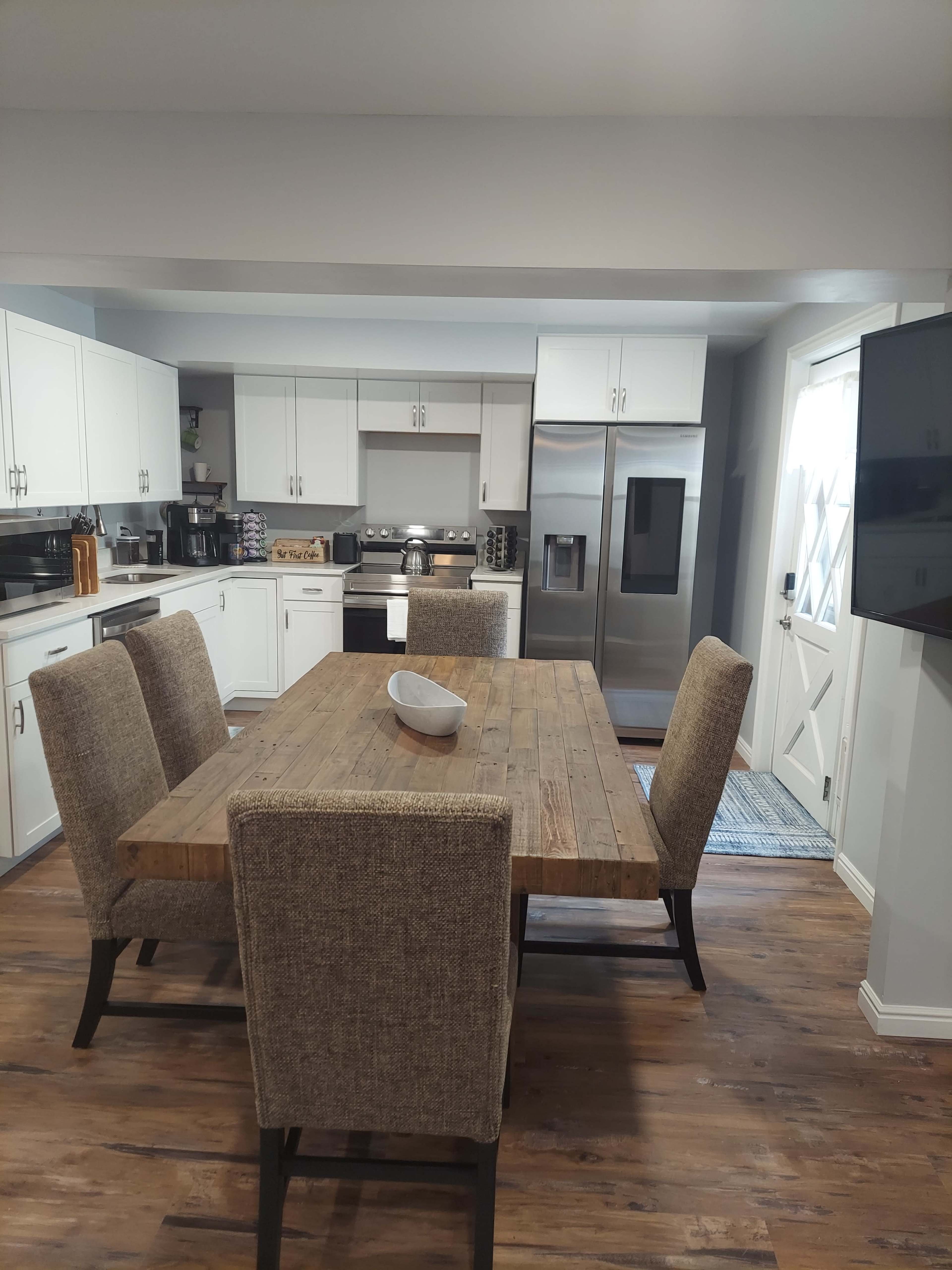 A modern kitchen with a large wooden dining table surrounded by upholstered chairs, stainless steel appliances, and white cabinetry.