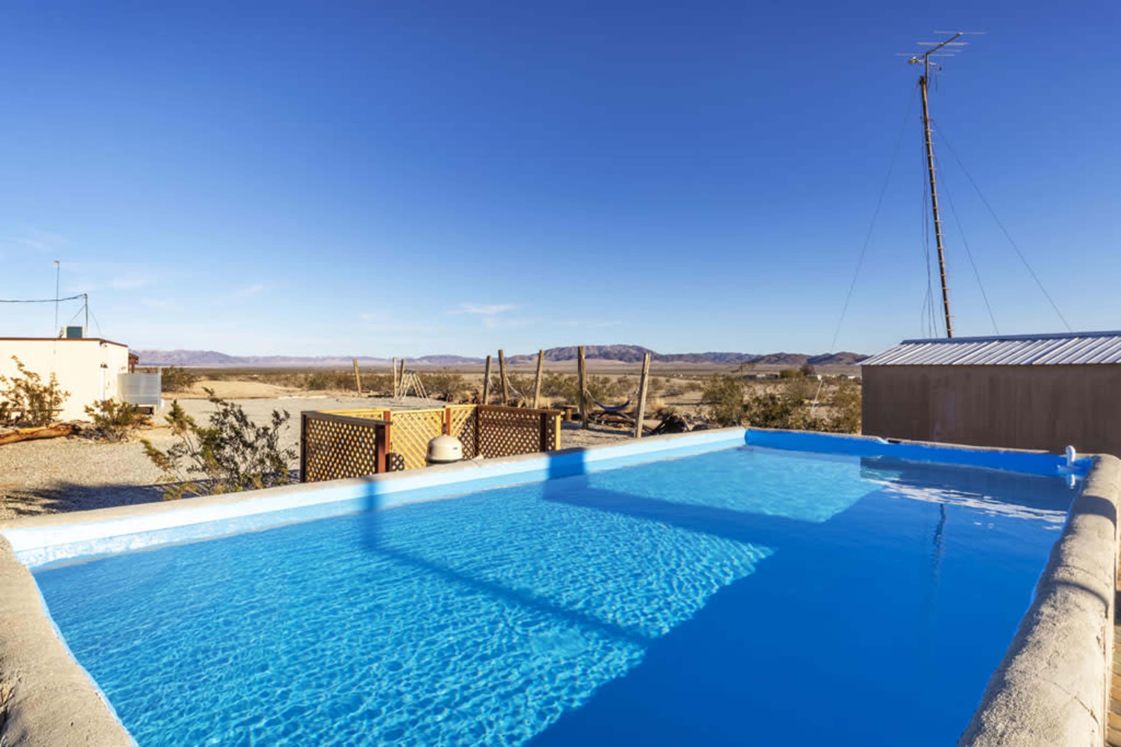 A rectangular swimming pool filled with clear blue water is set in a desert landscape, with mountains in the distance under a clear blue sky.