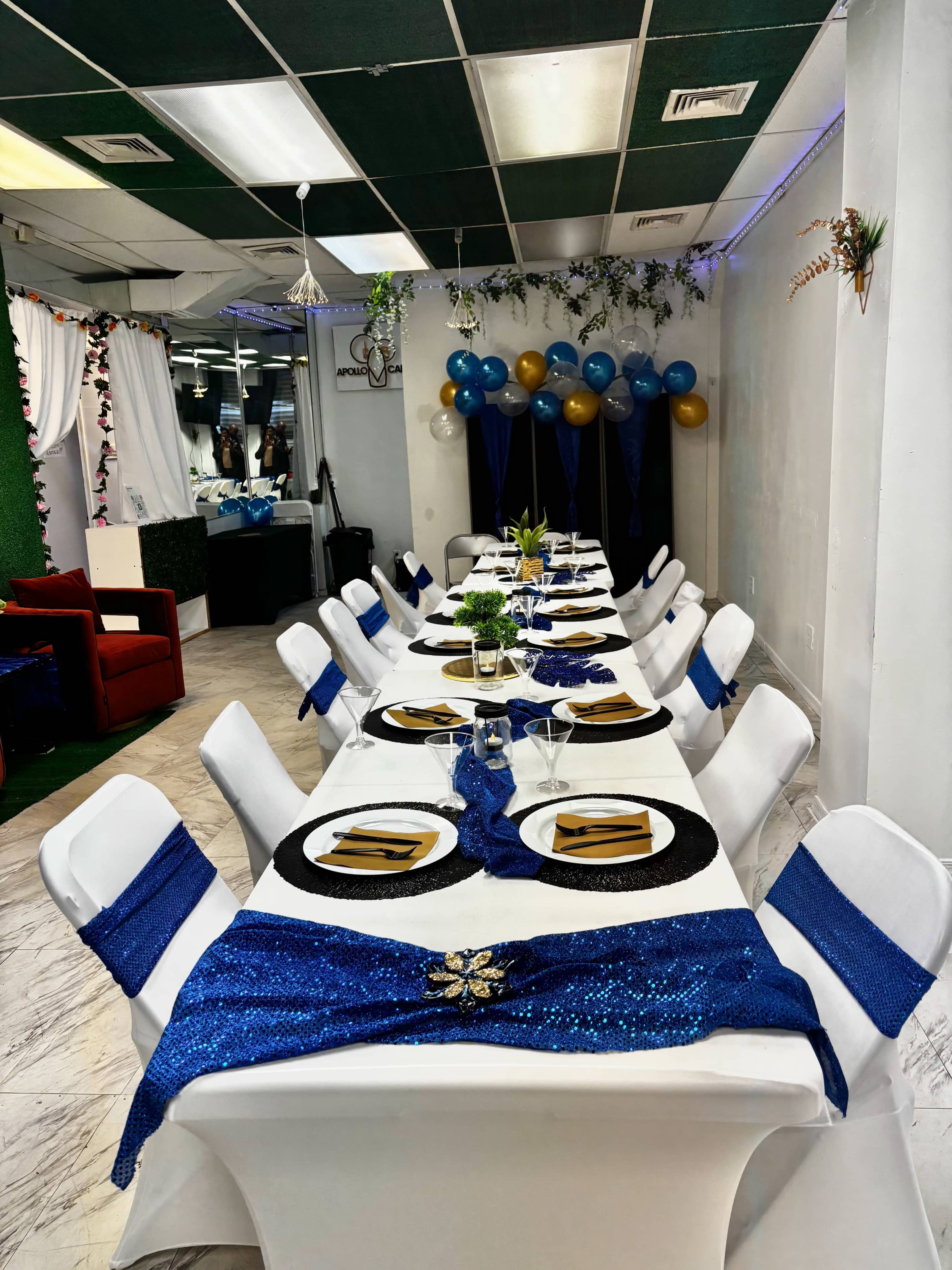 A long dining table is set with elegant tableware and decorated with blue and gold accents in a spacious room adorned with balloons and greenery.