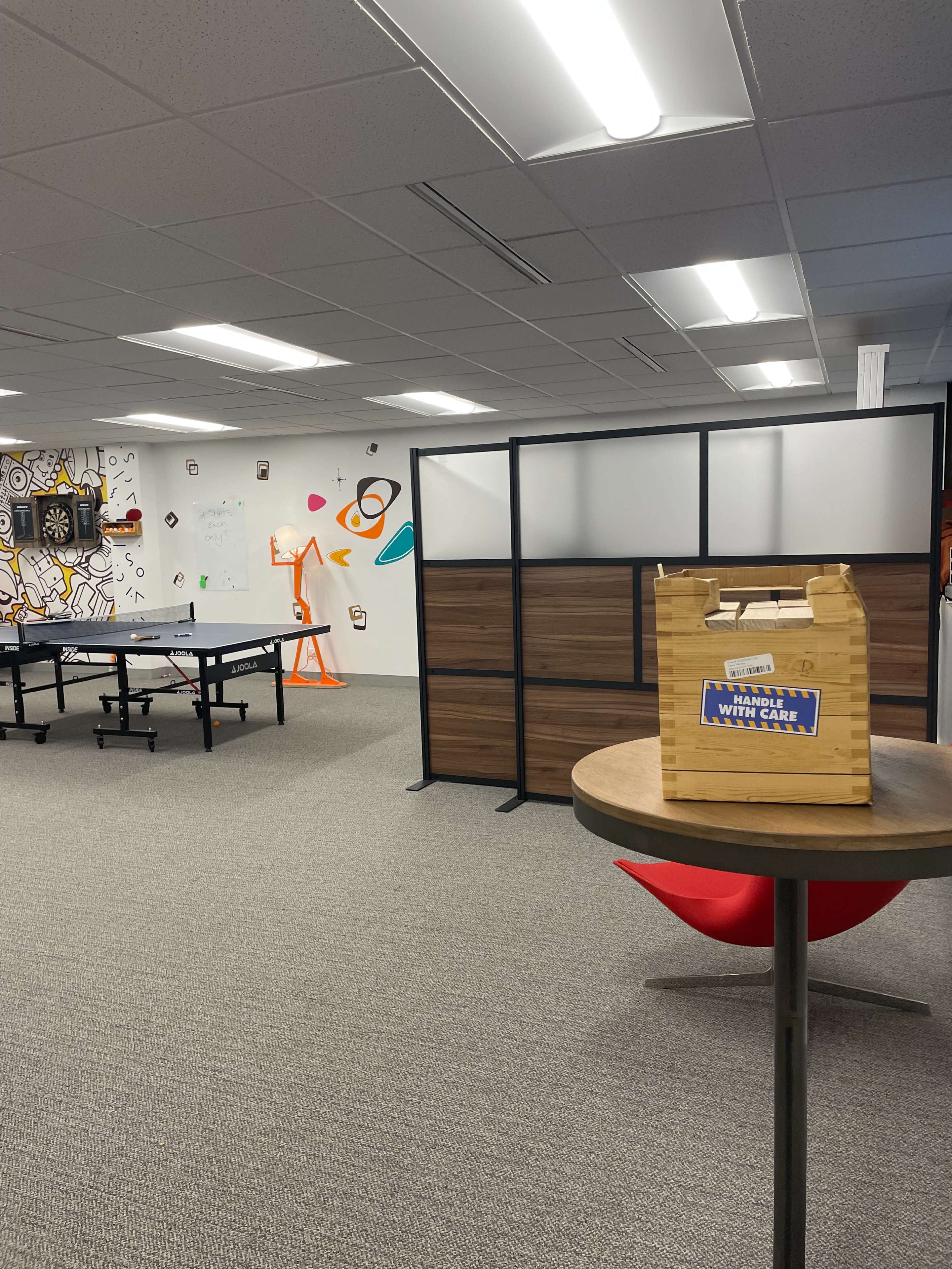 The image shows an office interior with a ping pong table, a partitioned area, and a circular table with a wooden box on it.