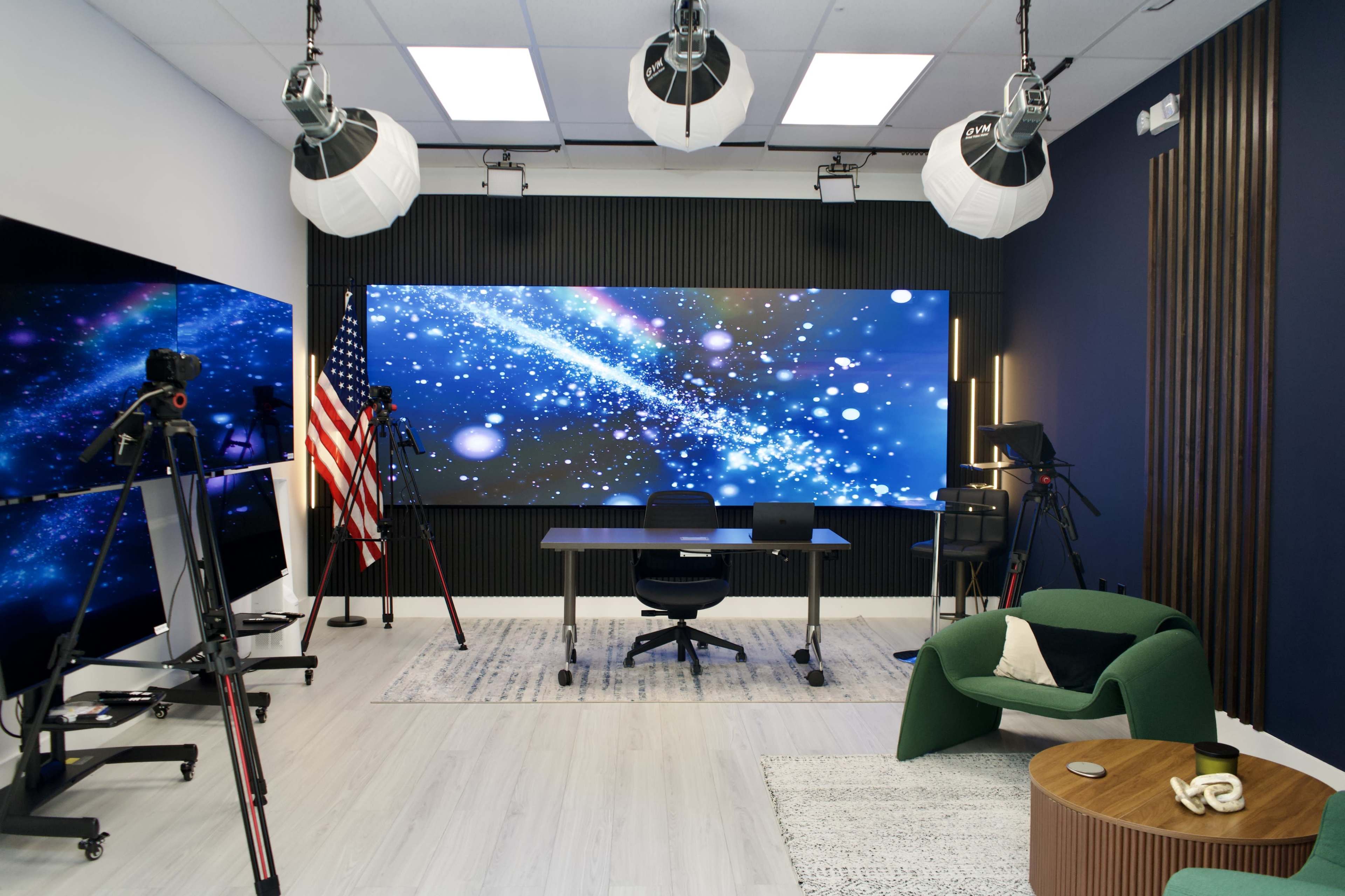 The image shows a modern studio setup featuring a large screen with a starry background, a desk with chairs, and various video equipment, alongside an American flag.