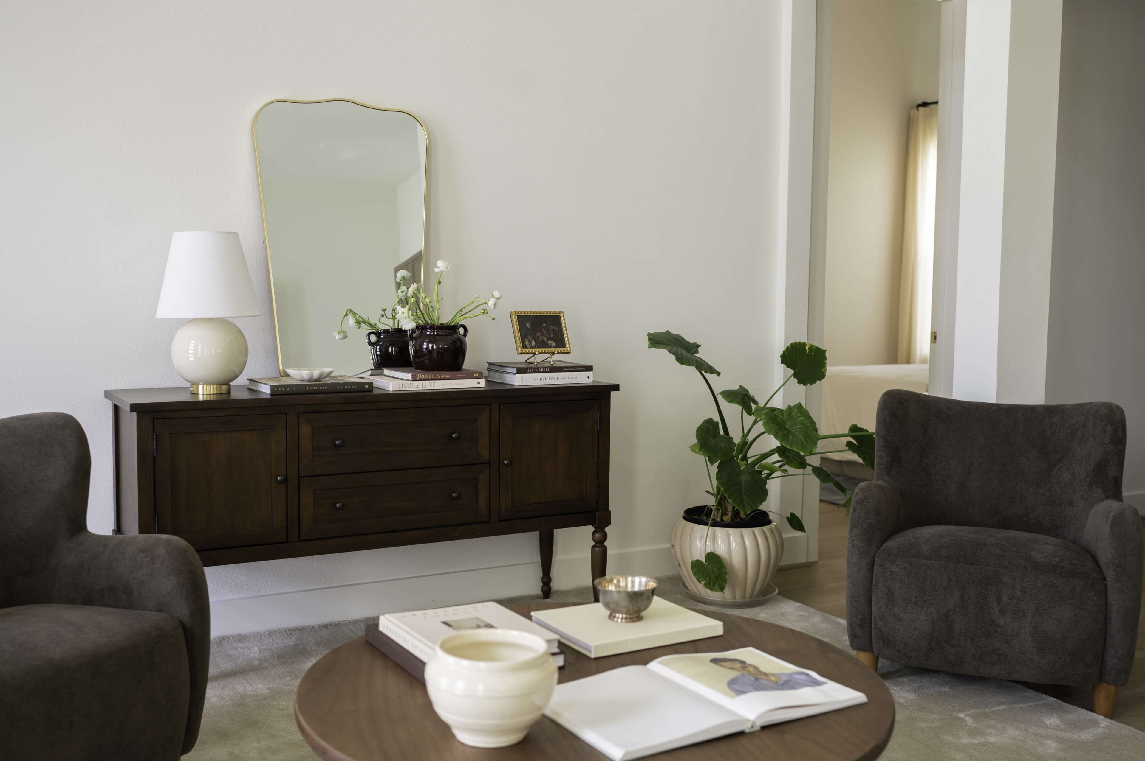 The image shows a cozy living room scene featuring a wooden sideboard, two upholstered chairs, and a coffee table adorned with books and decorative items.
