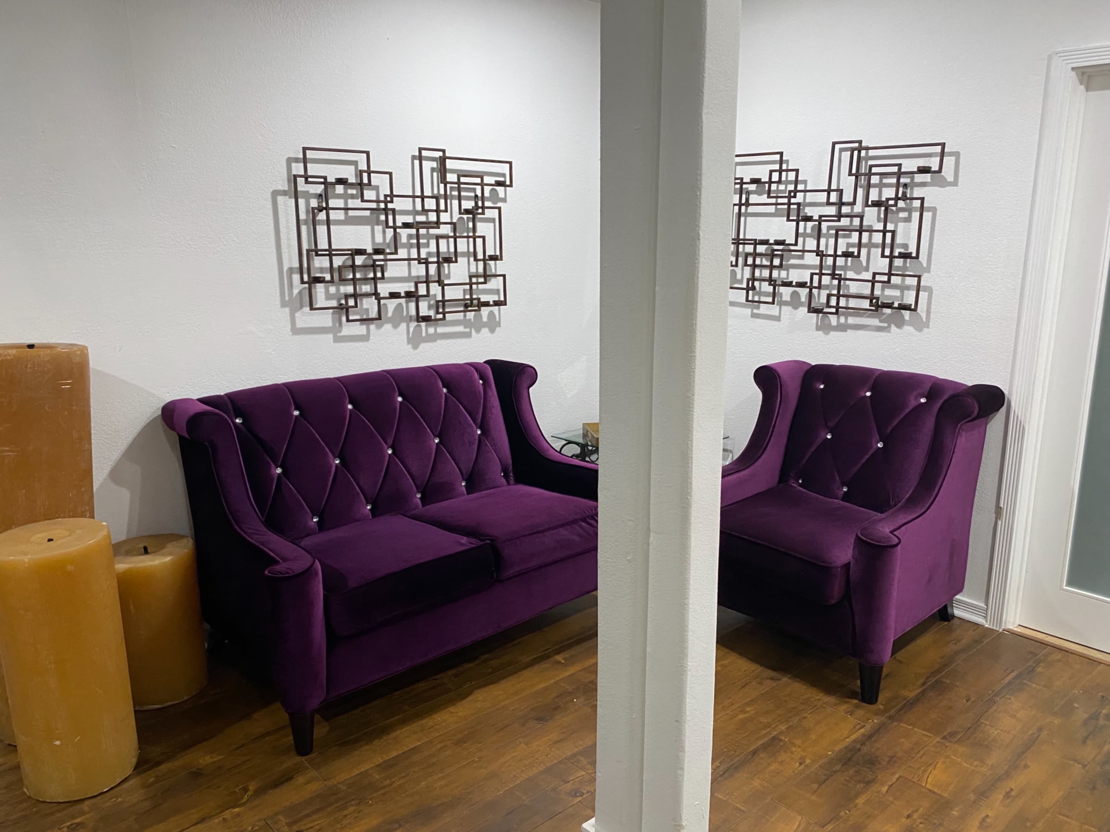 A pair of purple velvet sofas with tufted backs are positioned in a brightly lit room featuring modern wall sculptures.