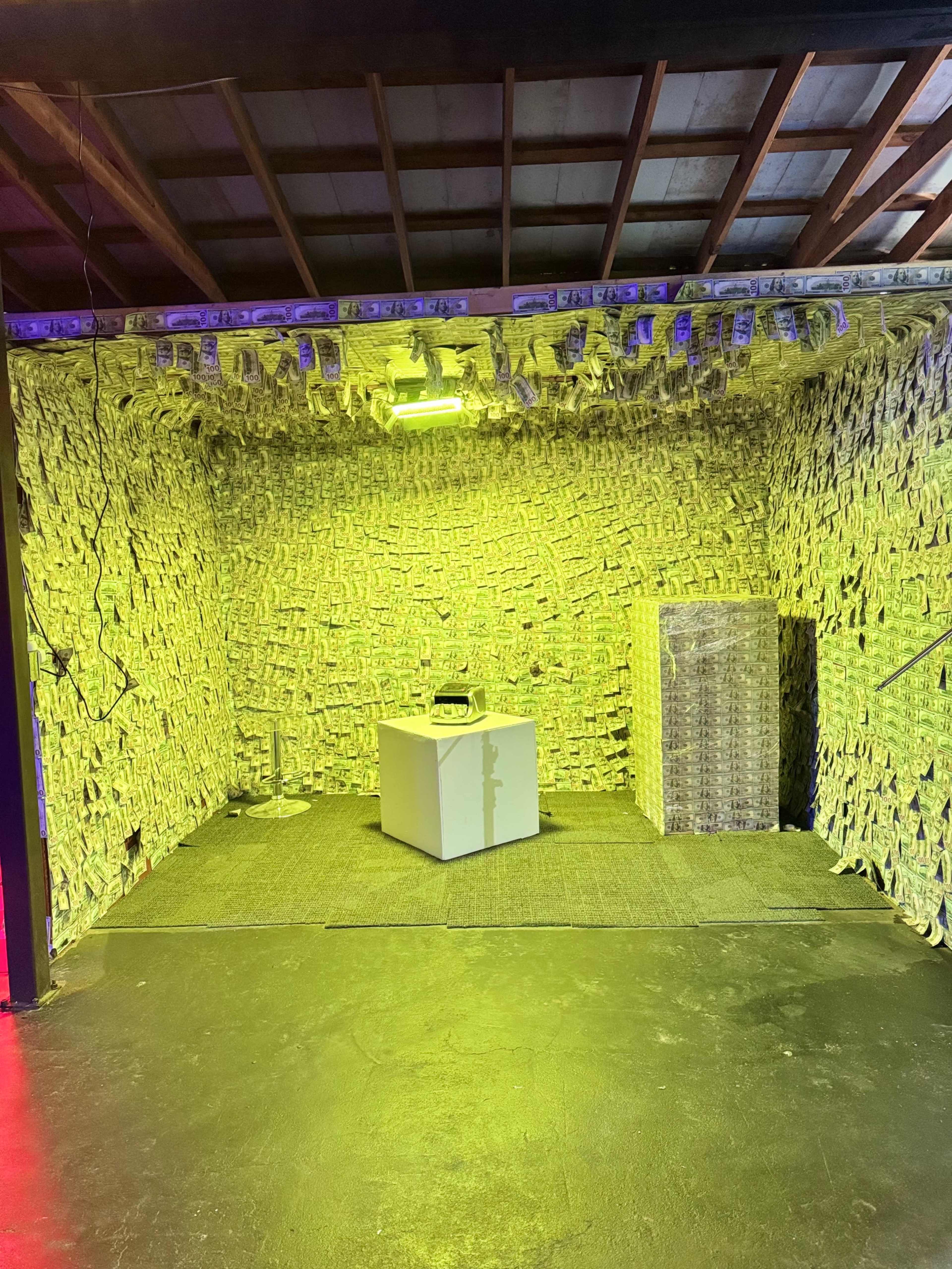 The image shows a room decorated with stacks of money covering the walls, featuring a white table and a stack of boxes in the center.