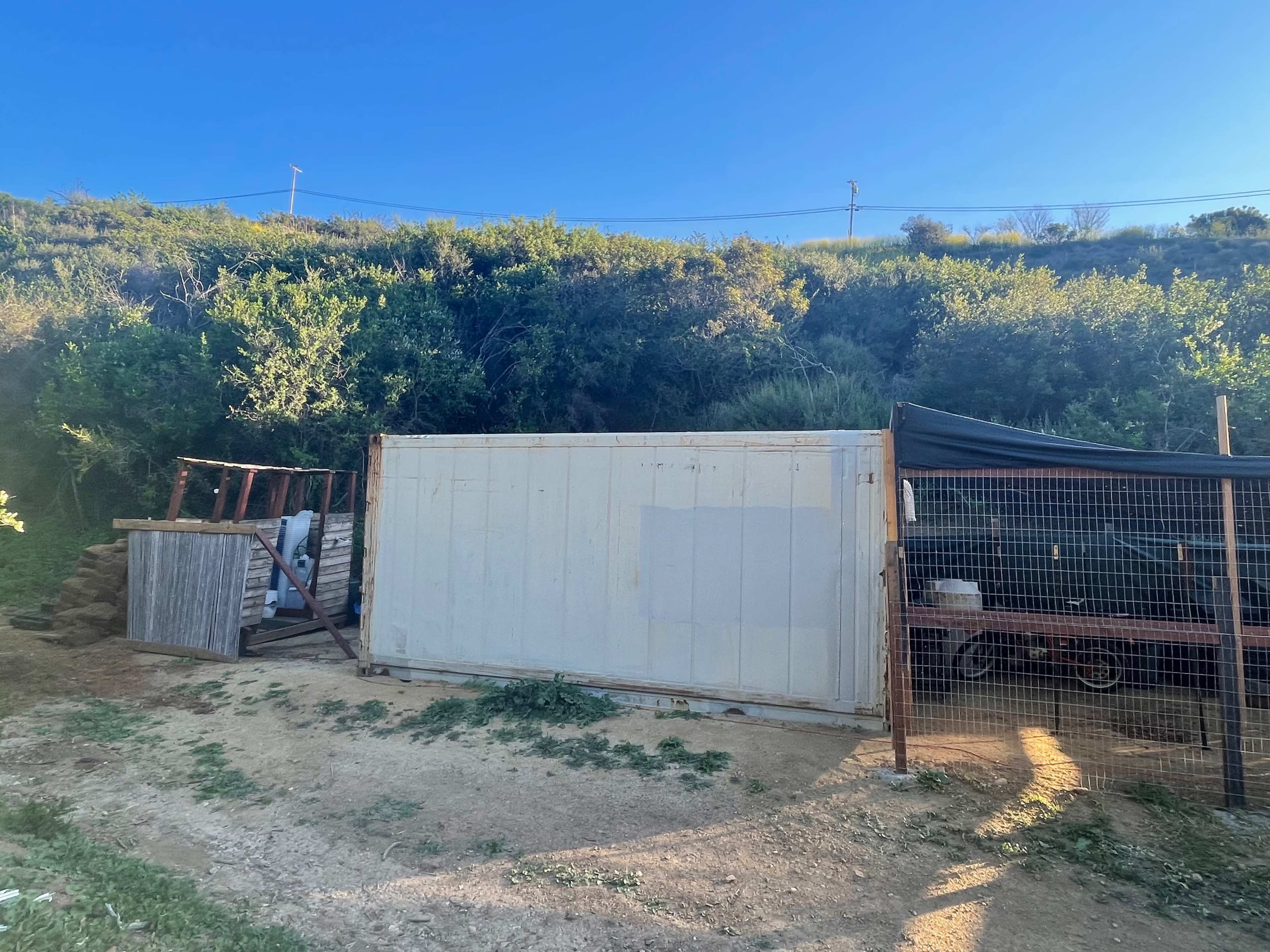 The image shows a white shipping container placed beside a fenced area on a grassy hillside.