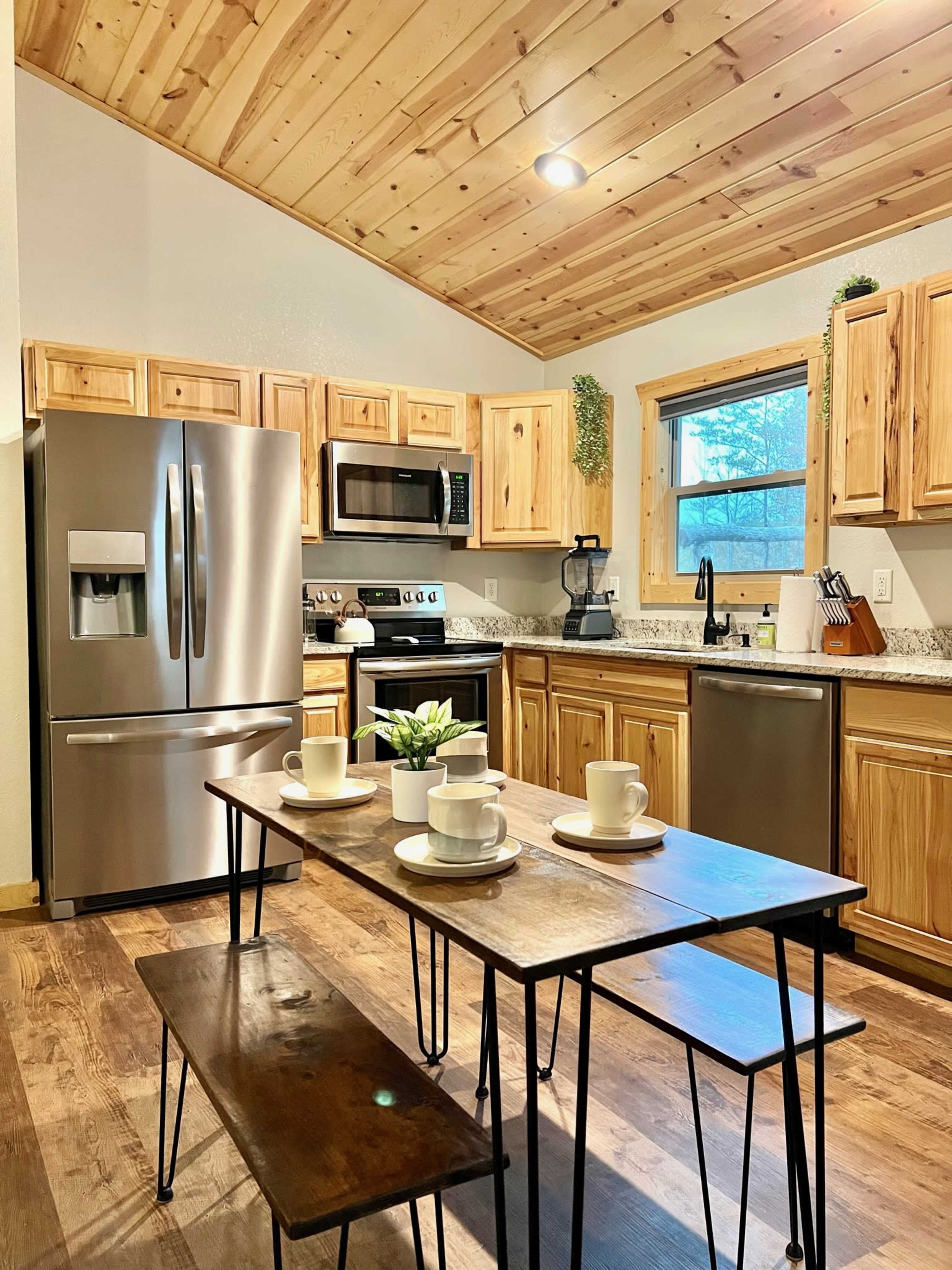 A kitchen features wooden cabinets, stainless steel appliances, and a dining table set with cups and a small plant.