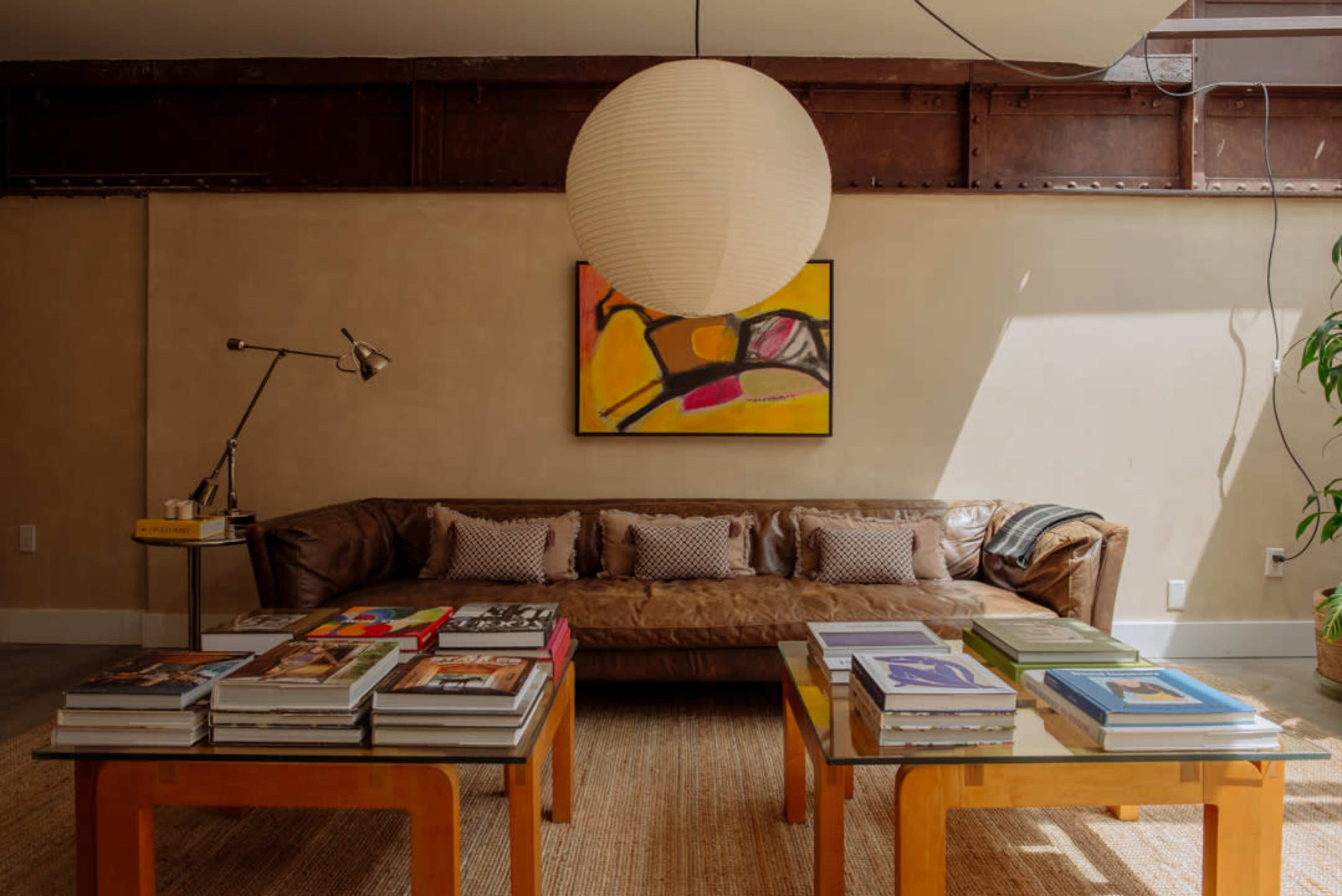 A cozy living room features a brown leather sofa, colorful artwork on the wall, and a coffee table stacked with books.
