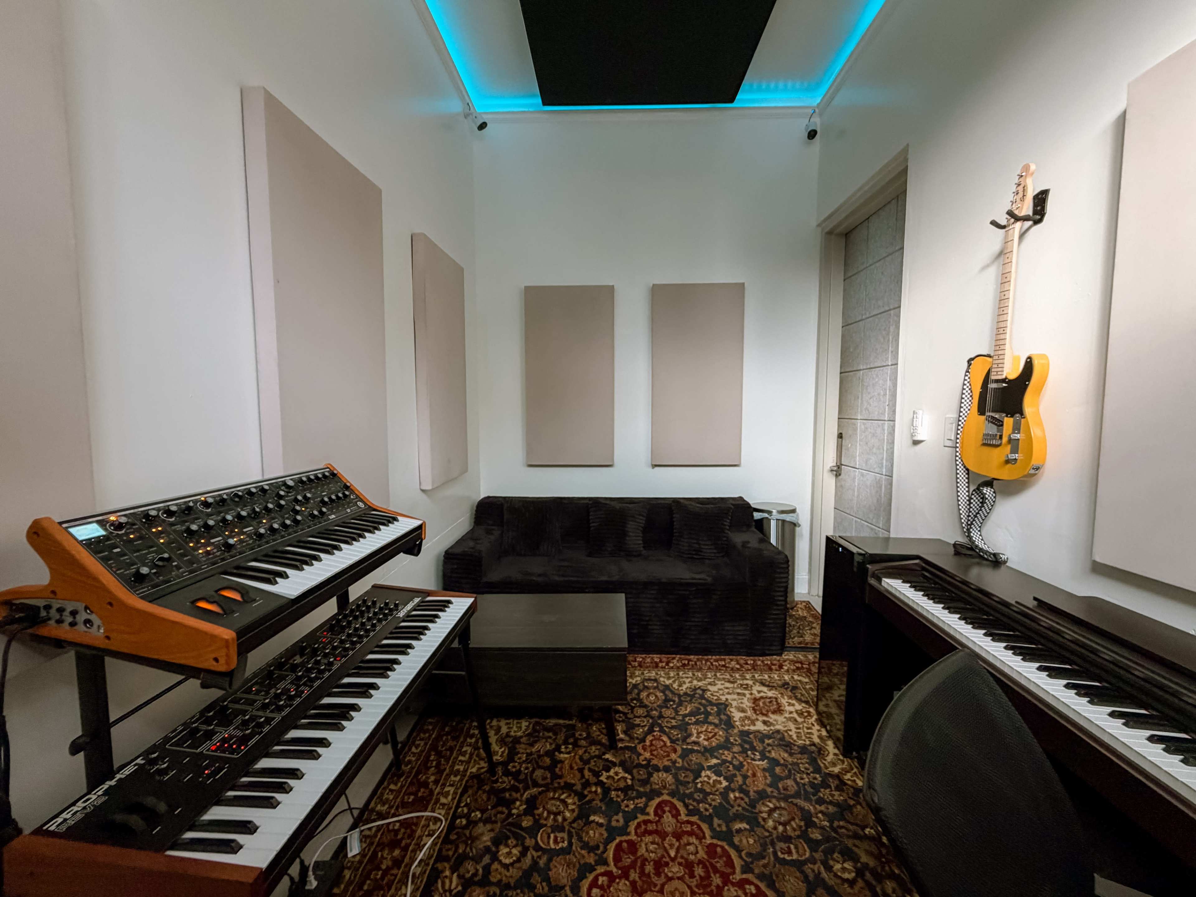 The image shows a music studio with synthesizers and a piano, a black couch, soundproof panels on the walls, and an electric guitar hanging above.