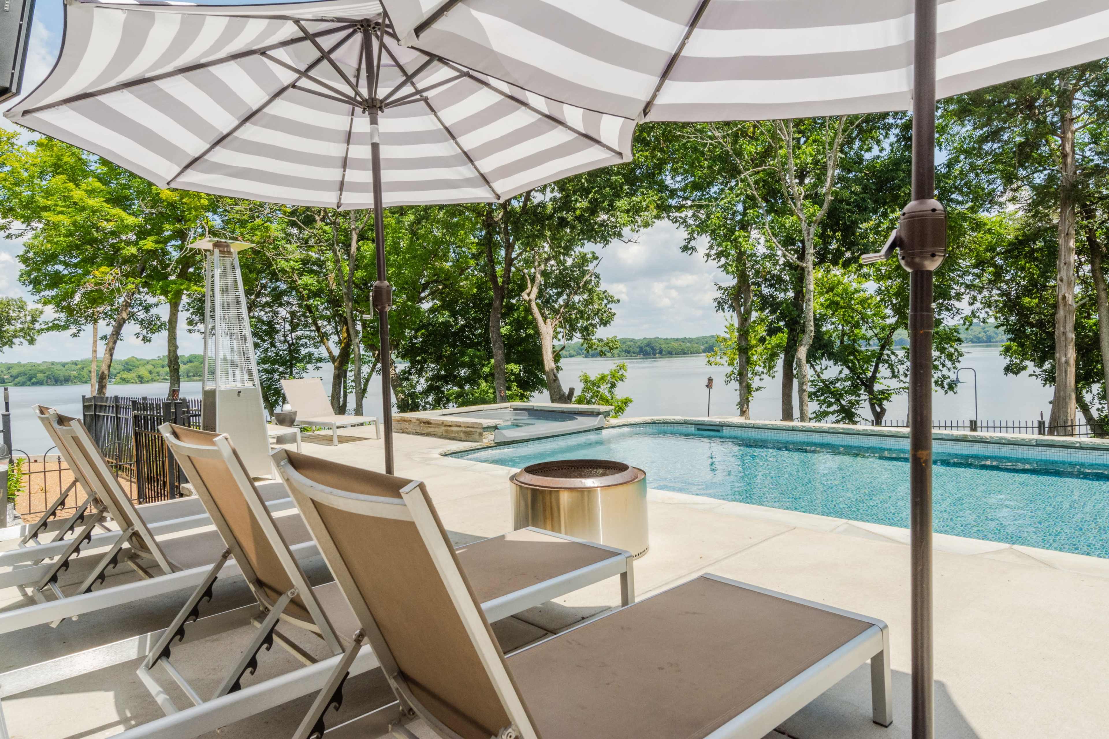 A pool area with lounge chairs and striped umbrellas overlooking a lake surrounded by trees.
