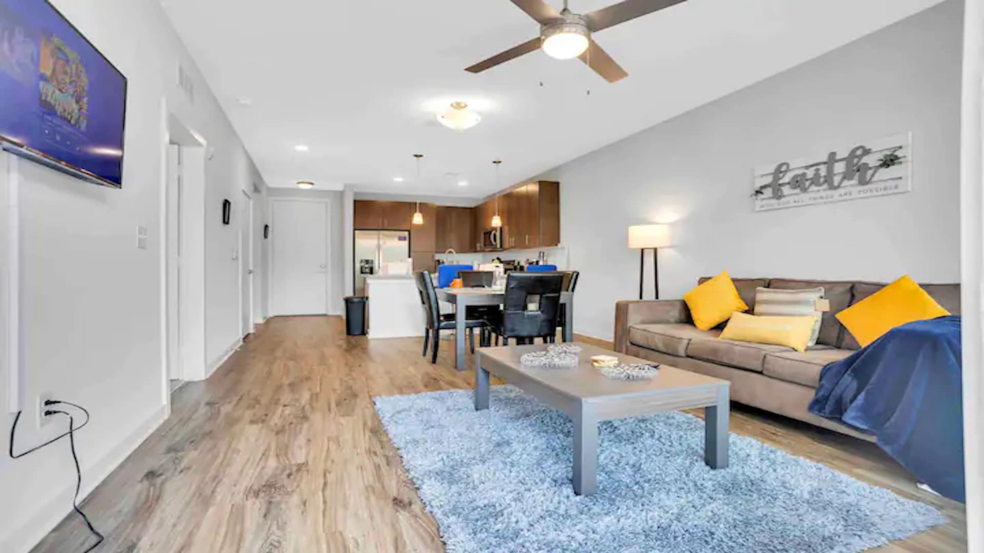 The image shows a modern living room with a brown sofa, a blue area rug, a dining table, and a kitchen in the background.