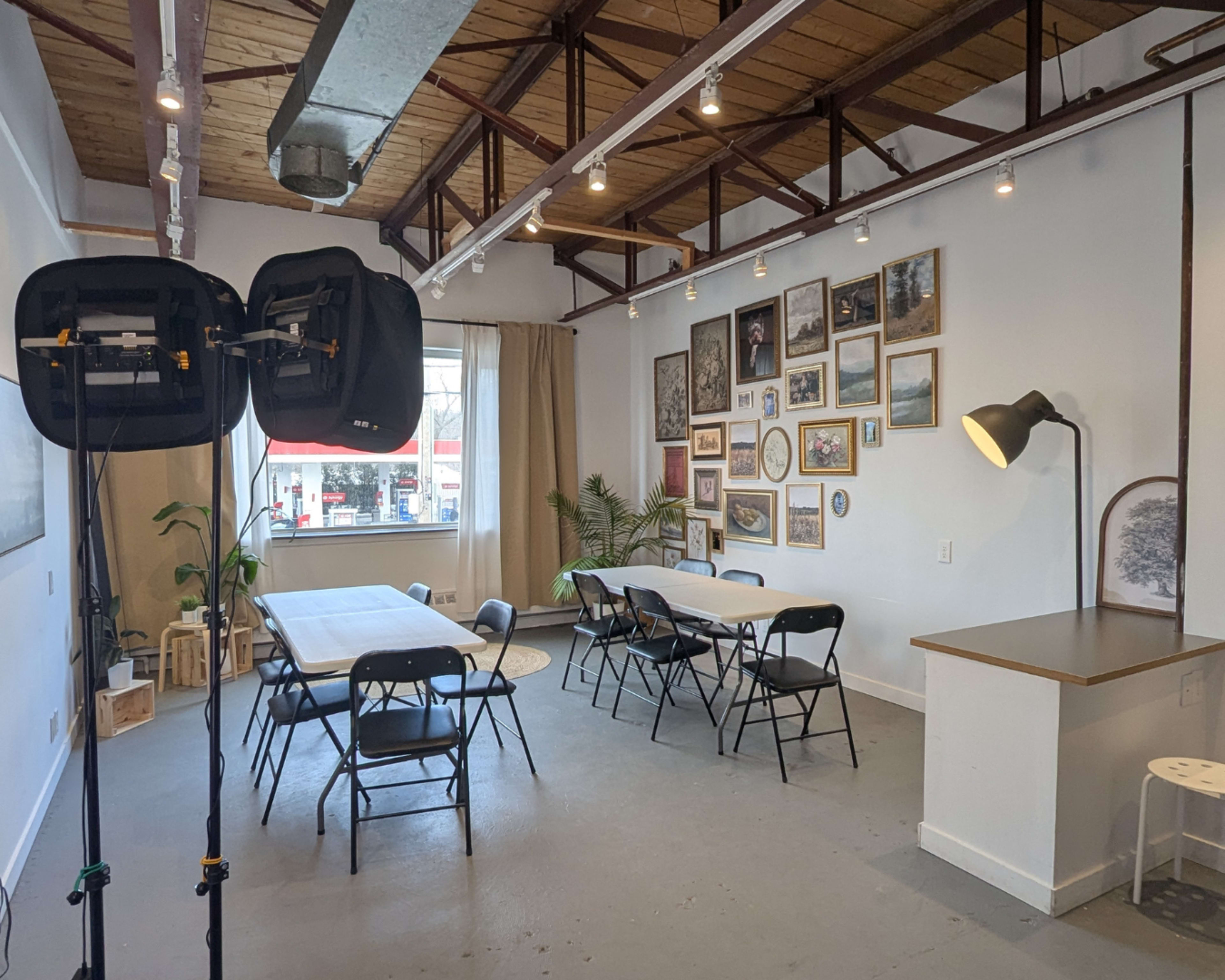 The image shows a bright, open room with several tables and chairs arranged for a meeting, along with a backdrop of framed artwork on the walls.