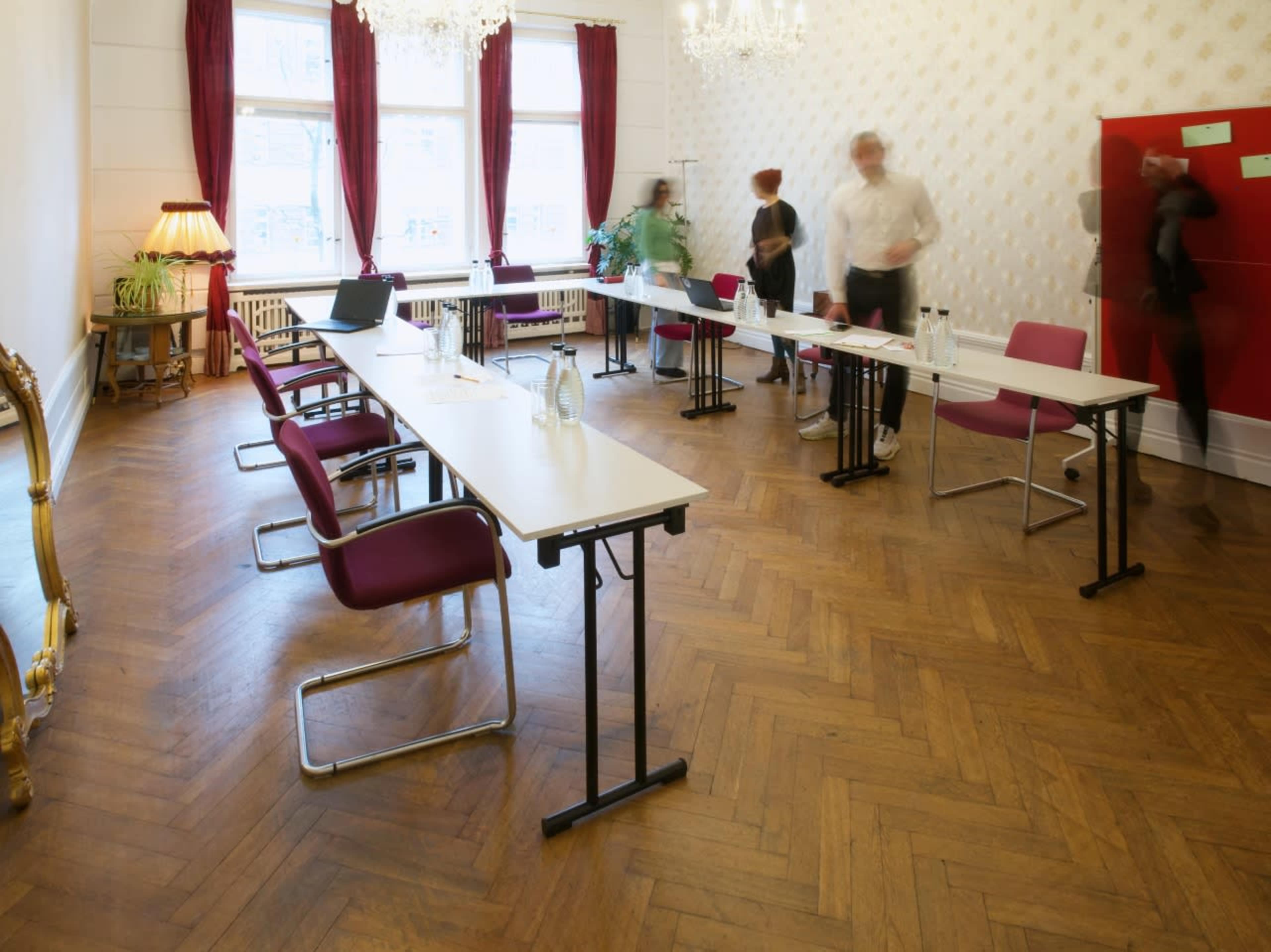 A meeting room with several tables and chairs arranged, featuring a window with red curtains and a chandelier overhead.