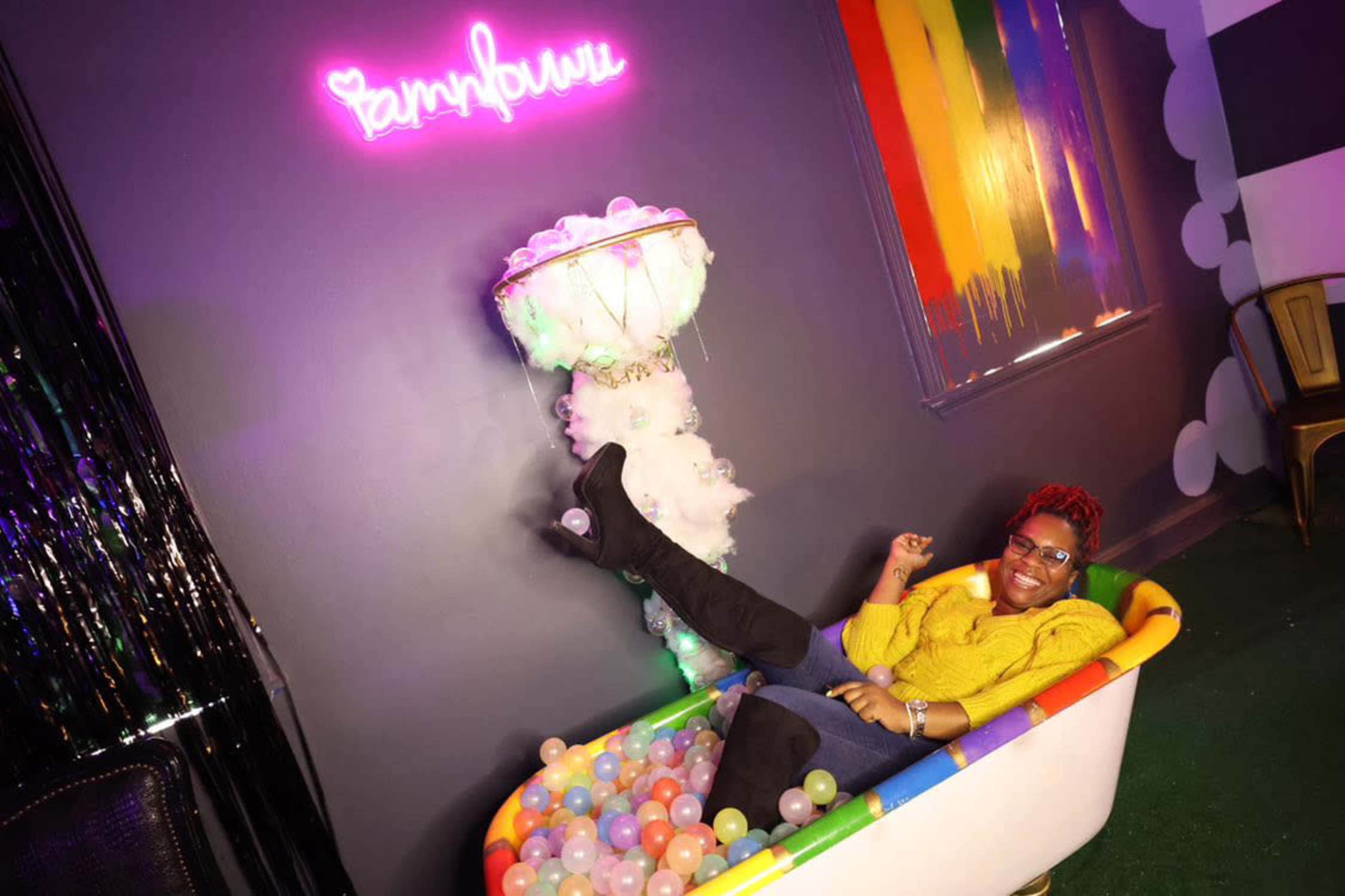 A person is sitting in a bathtub filled with colorful balls, with a neon sign above that reads "sampbulu" and a rainbow-striped wall in the background.