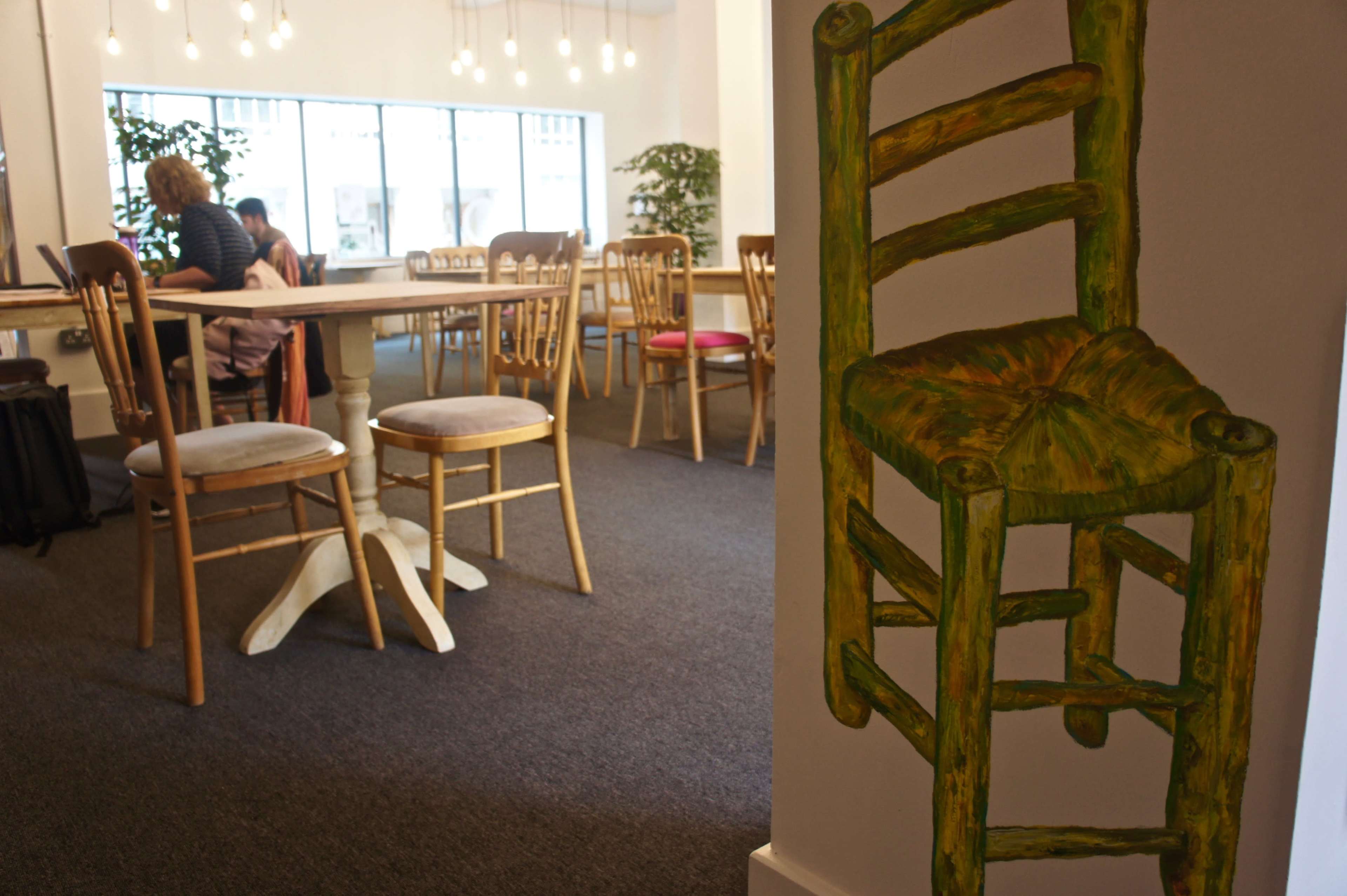A spacious, modern café scene with wooden chairs, a large table, and a painted green chair on the wall.