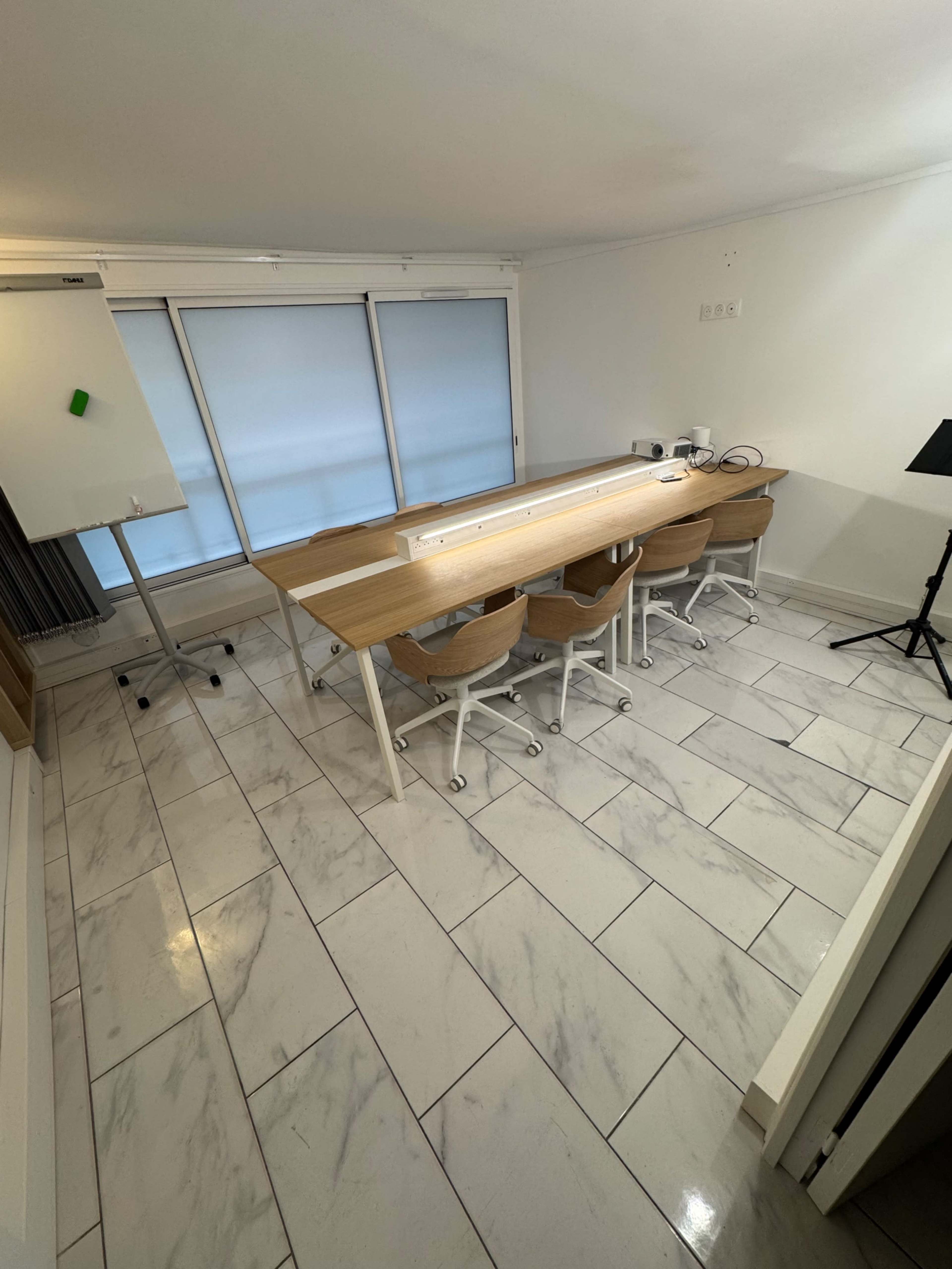 The image shows a modern meeting room featuring a long table, several chairs, a whiteboard, and large windows with blinds.