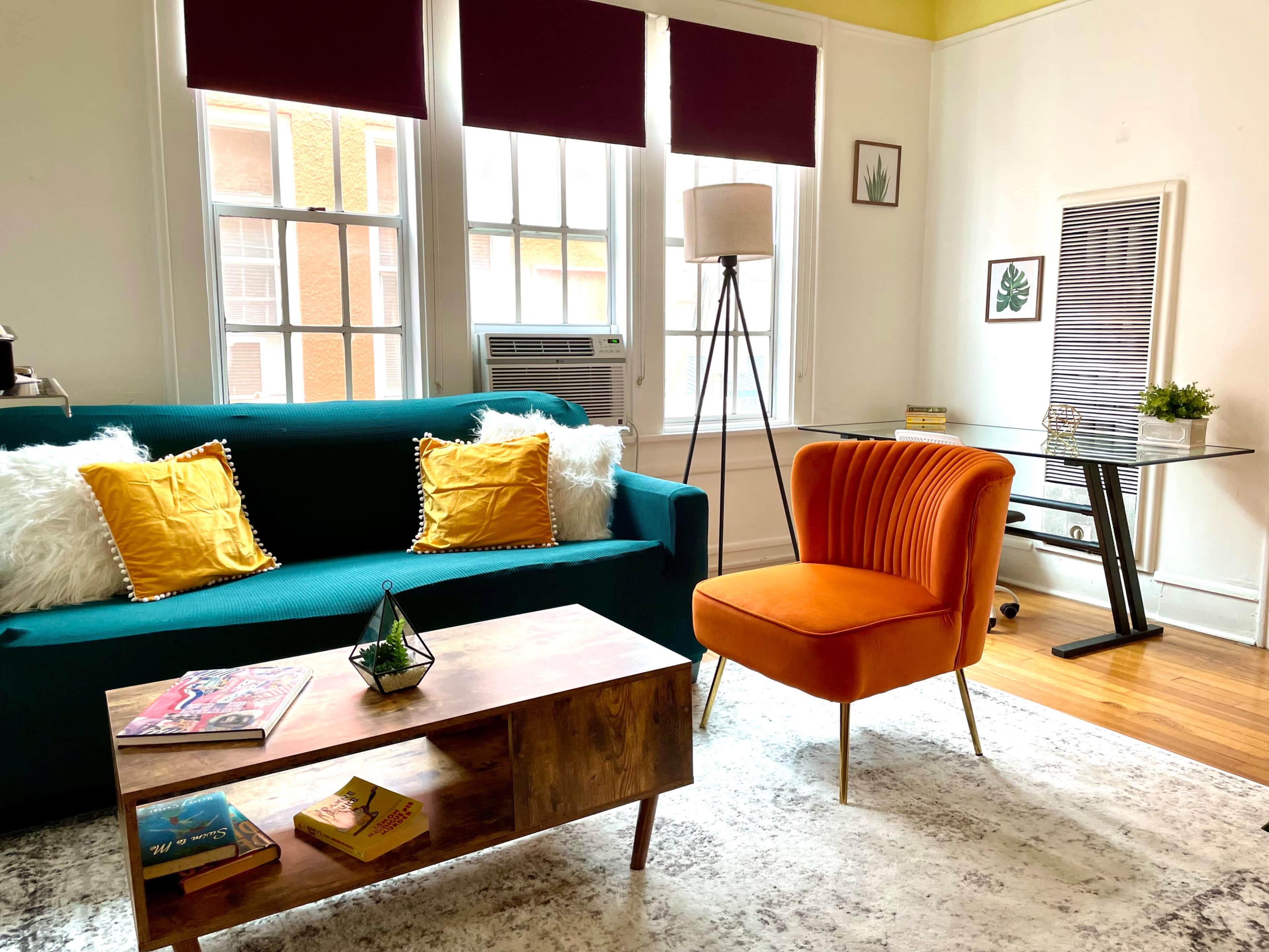 A cozy living room with a teal sofa, an orange accent chair, a wooden coffee table, and large windows letting in natural light.