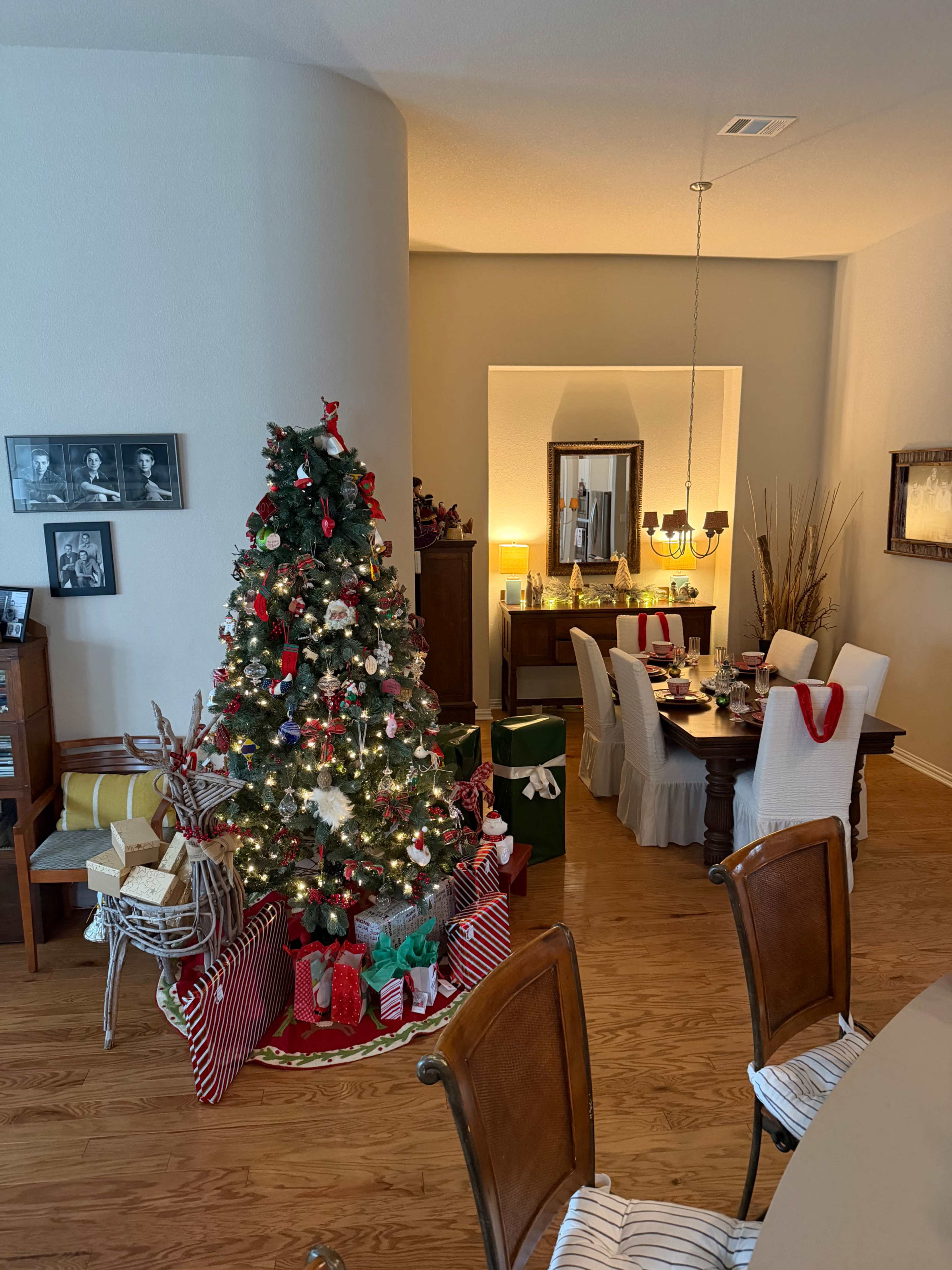 A decorated Christmas tree with presents is positioned next to a dining area set for a festive meal in a brightly lit room.