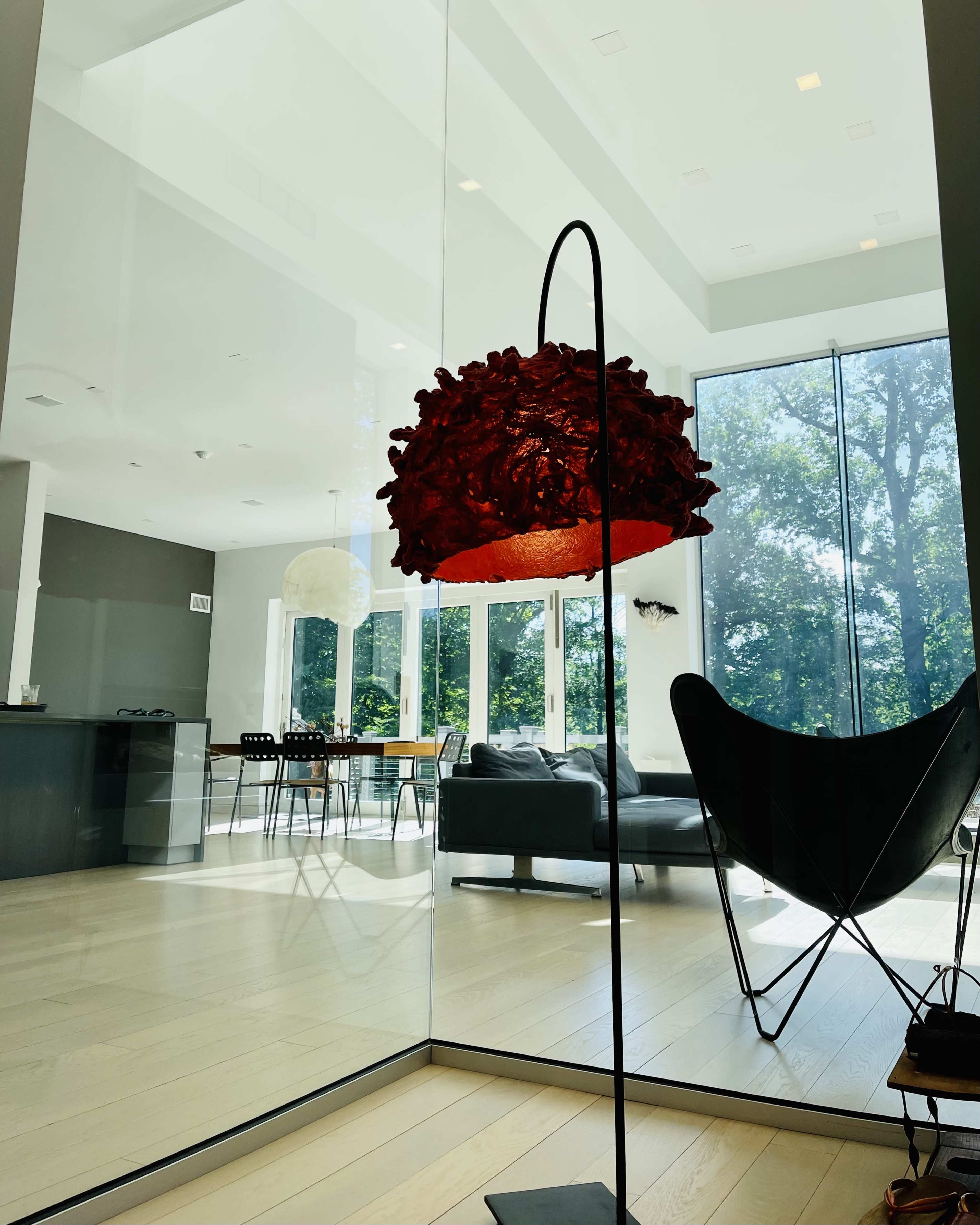 A modern interior features a decorative red lamp in the foreground, with a dining area and large windows in the background.