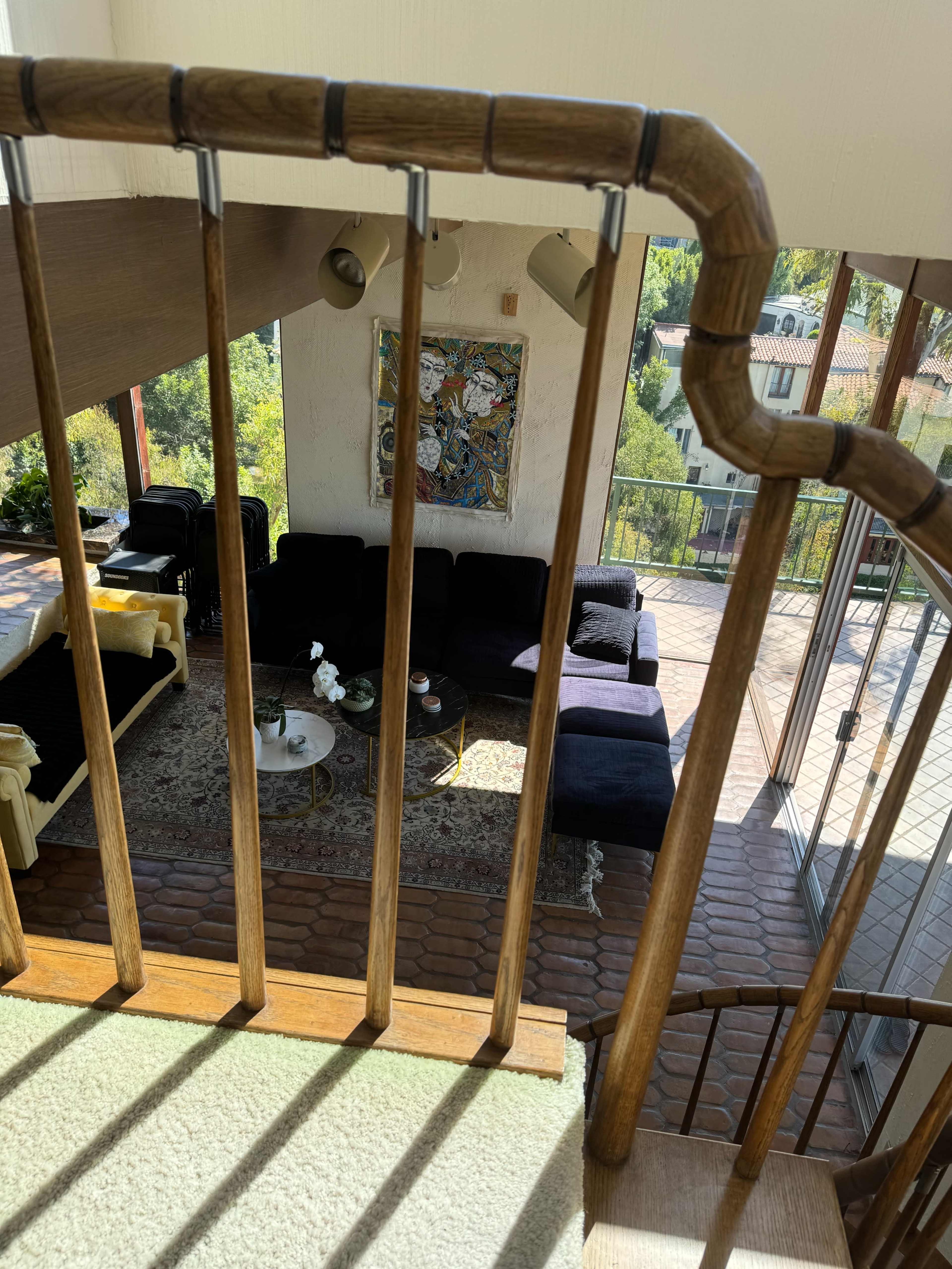 A view from a staircase captures a sunlit living room featuring a dark sectional sofa, a coffee table, and large windows overlooking a green landscape.