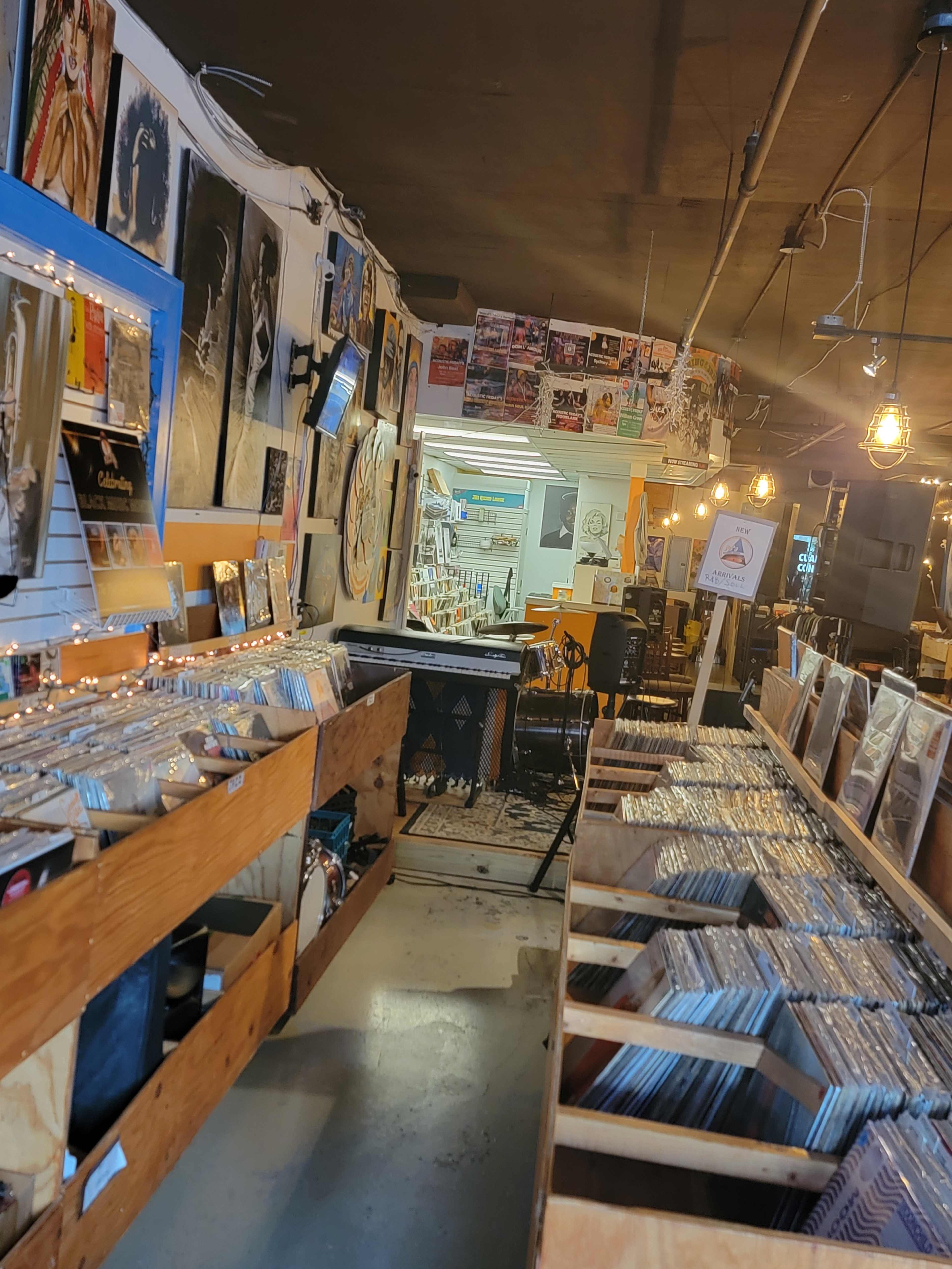 The image shows the interior of a record store with rows of wooden crates filled with vinyl records and posters covering the walls.
