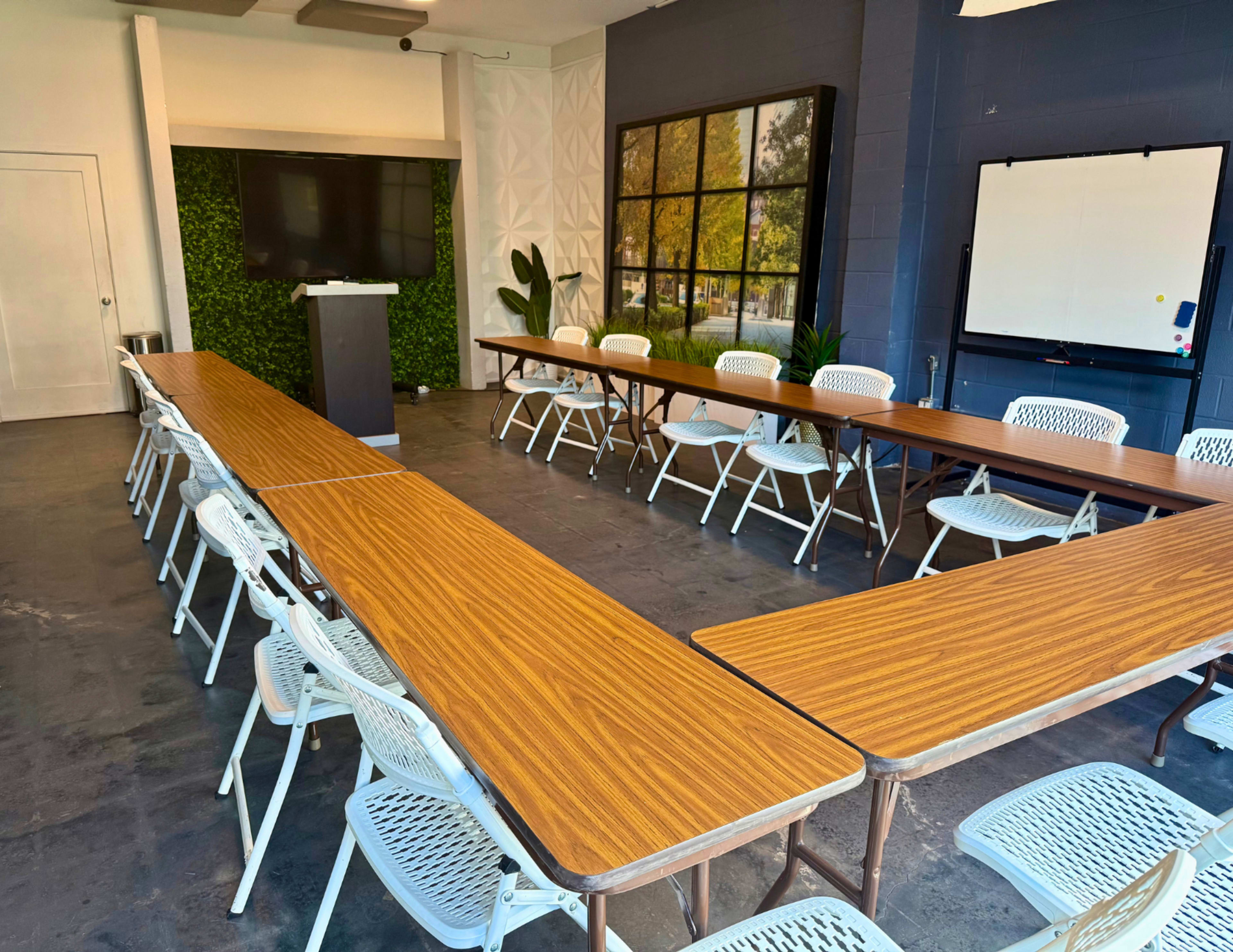 A large meeting room features multiple arranged tables with white chairs, a television screen, and green wall foliage.