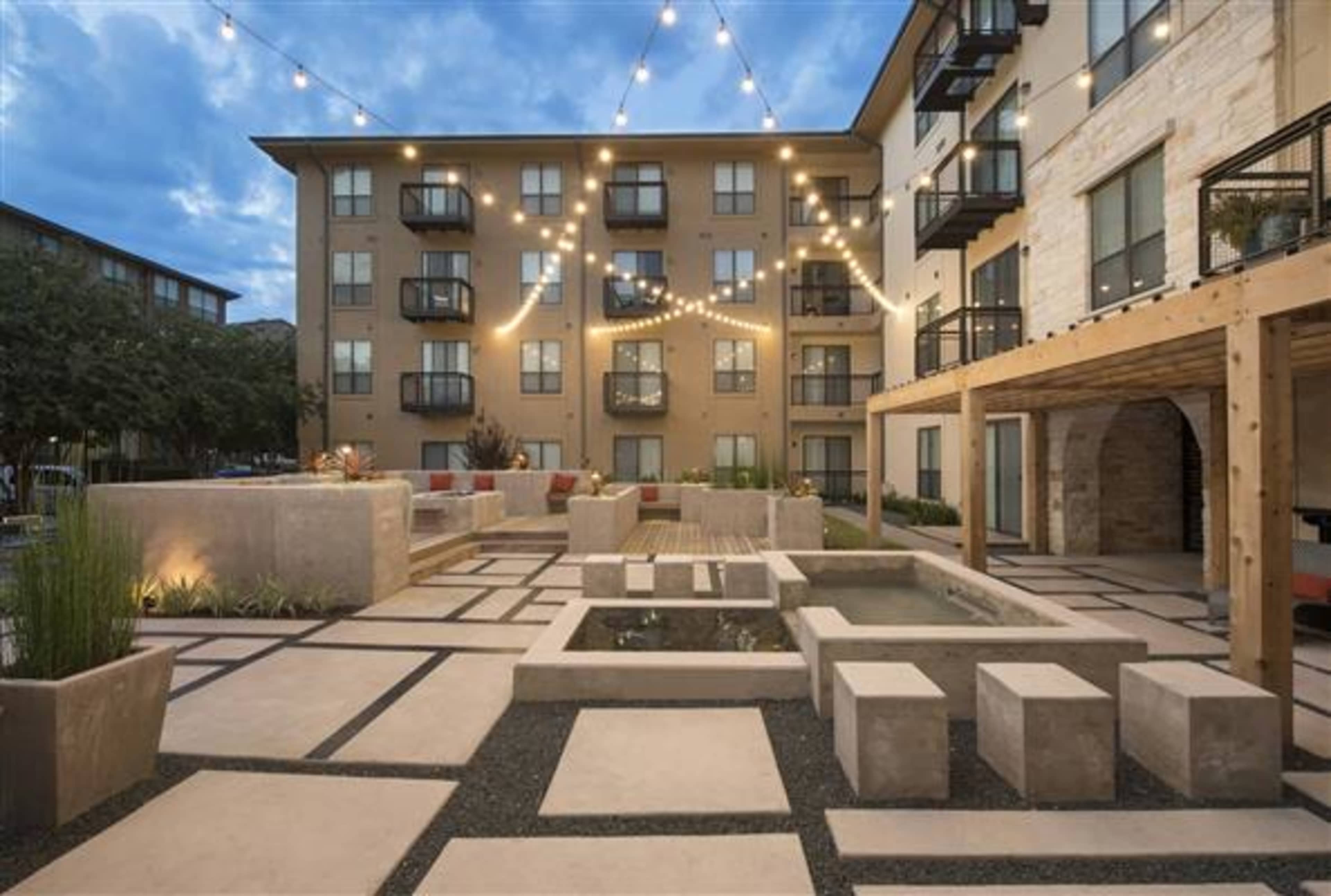 The image shows a modern outdoor courtyard featuring geometric stone pathways, planters, and a water feature, surrounded by apartment buildings and decorative string lights.