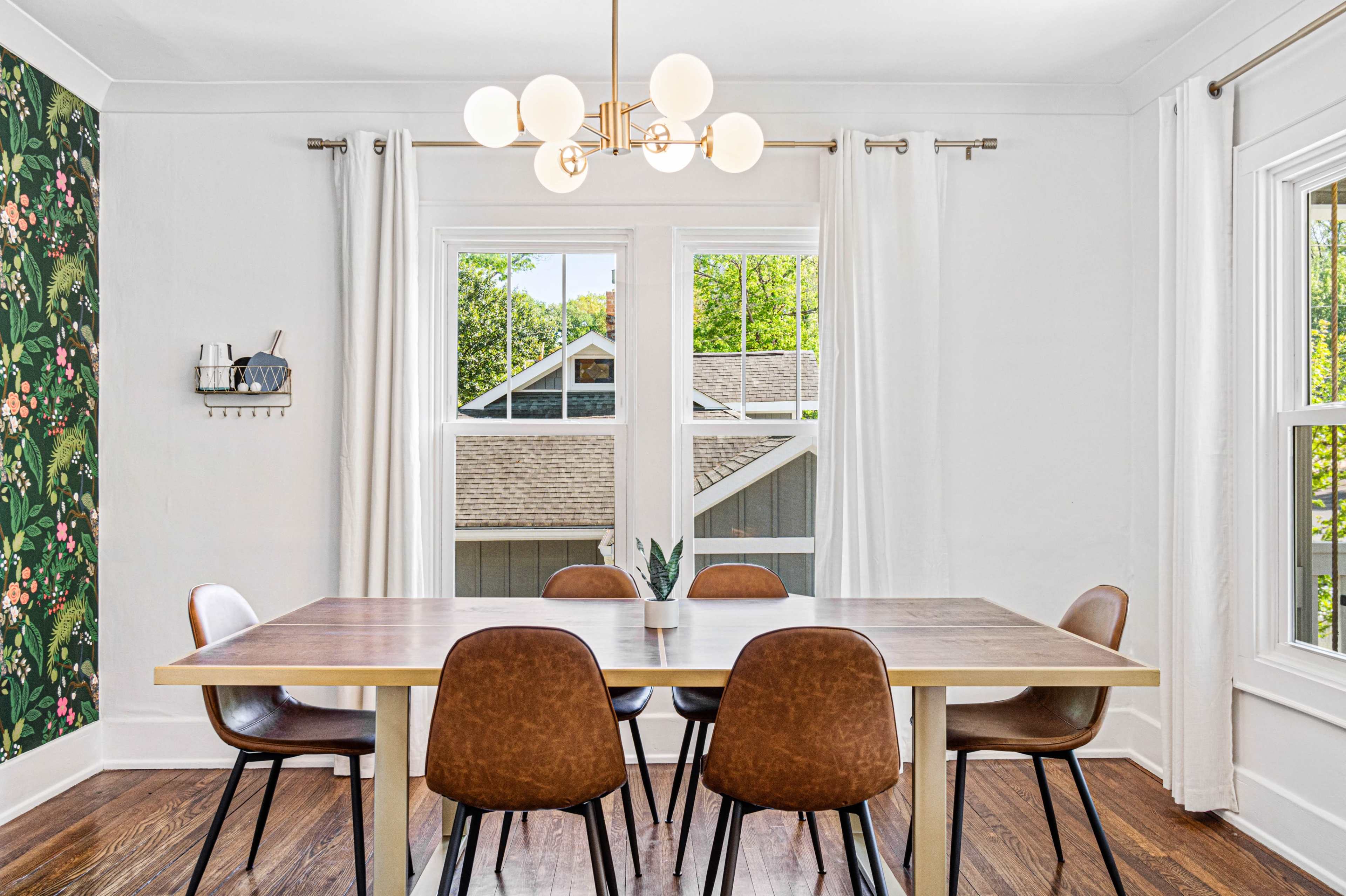 A dining room features a wooden table surrounded by brown chairs, with large windows and floral wallpaper on one wall.