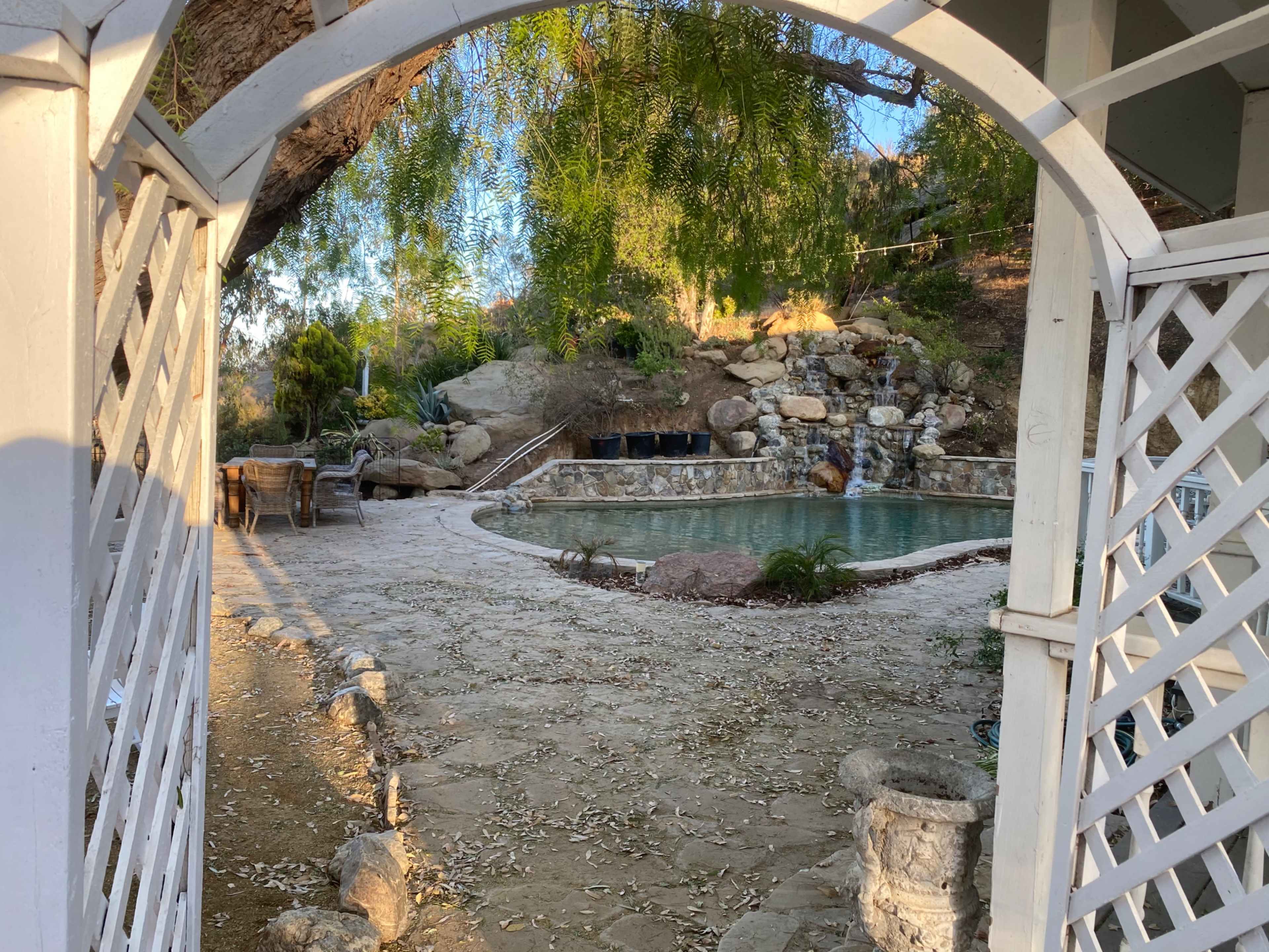 A stone-paved area with a swimming pool surrounded by rocks and greenery, framed by a white archway.
