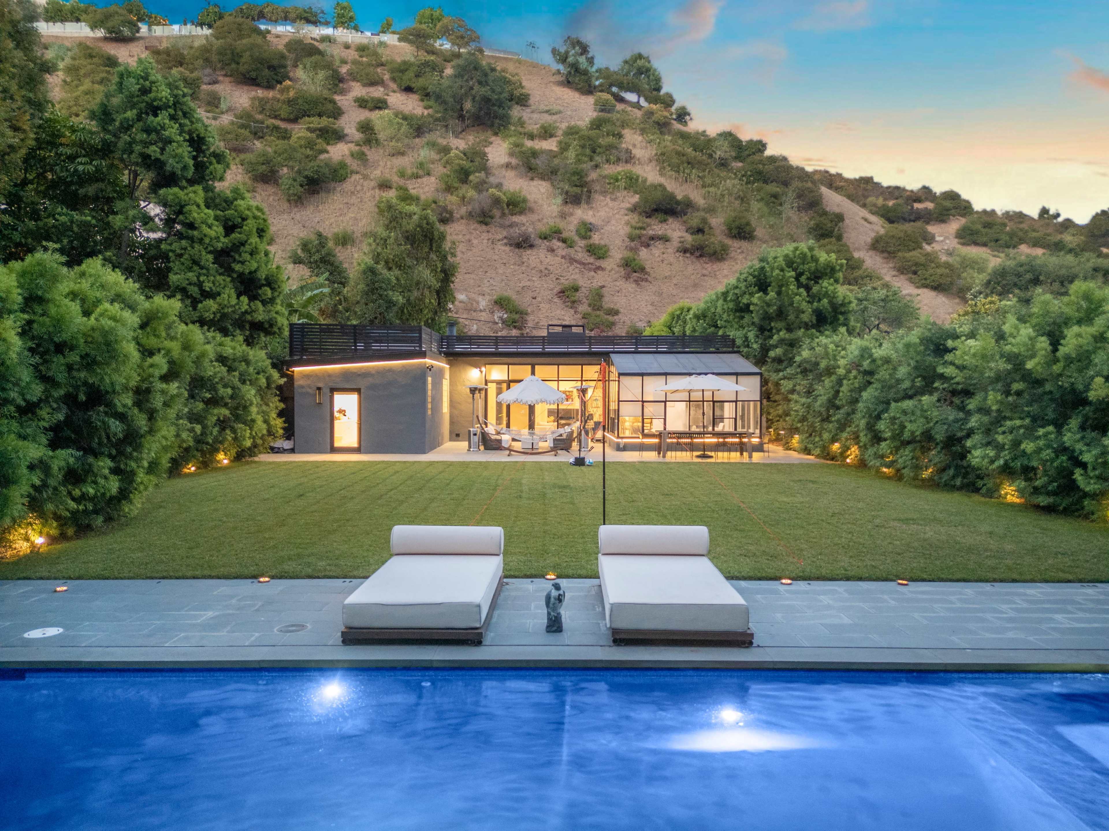 The image shows a modern house set against a hillside, with a pool in the foreground and a well-lit lawn leading to a patio shelter.