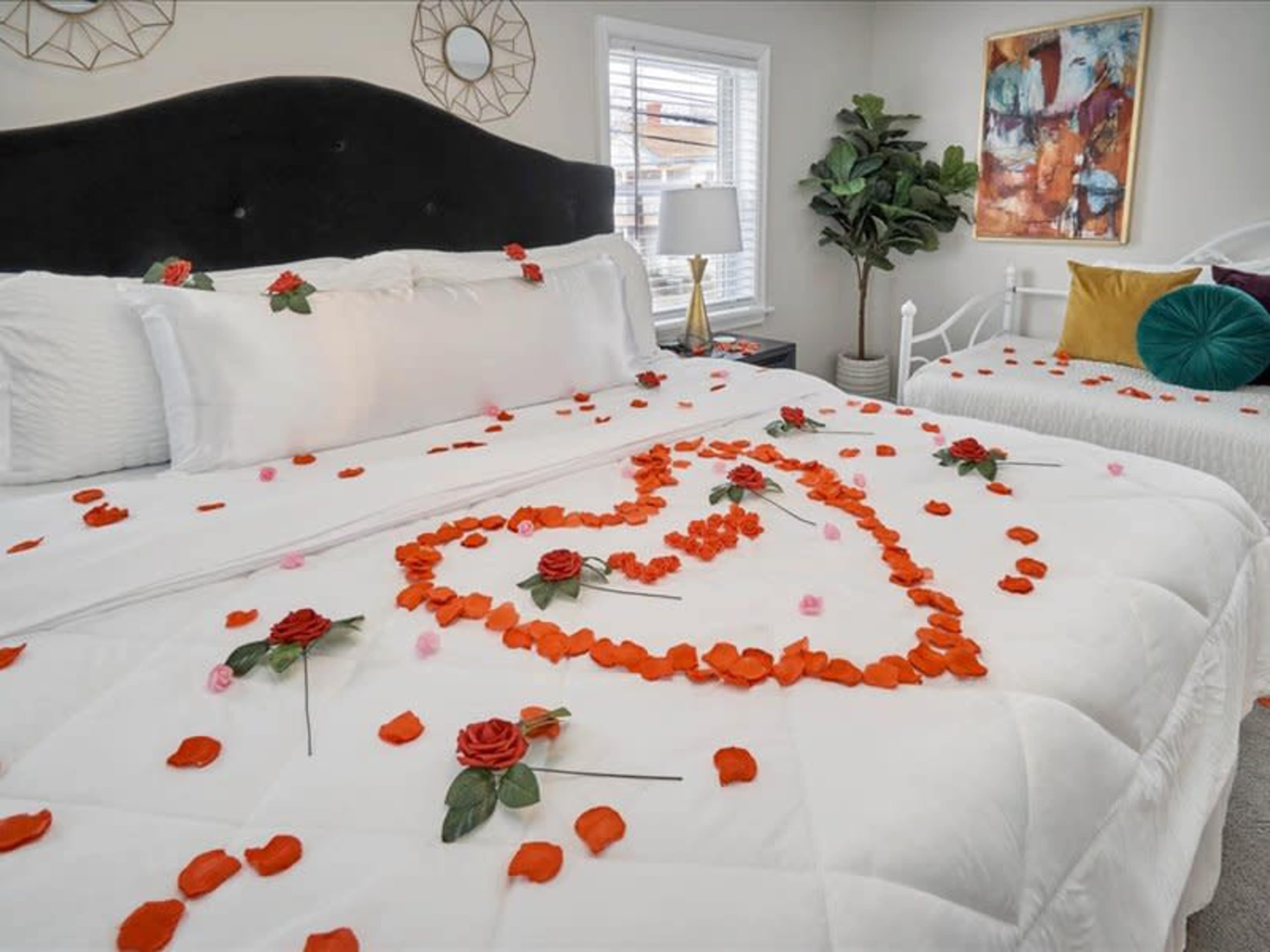 A bed is adorned with rose petals arranged in a heart shape, surrounded by additional floral decorations in a neatly arranged room.